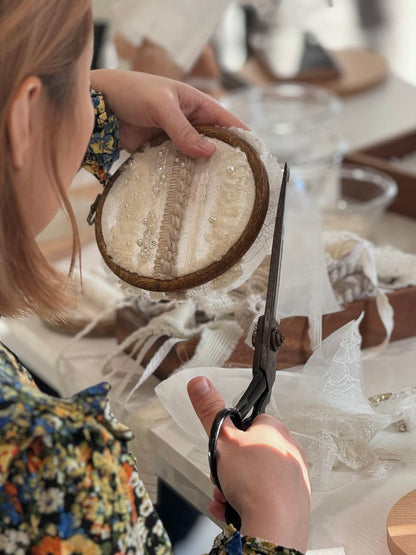 Person holding a small embroidery hoop with lace and scissors in a blurred indoor setting