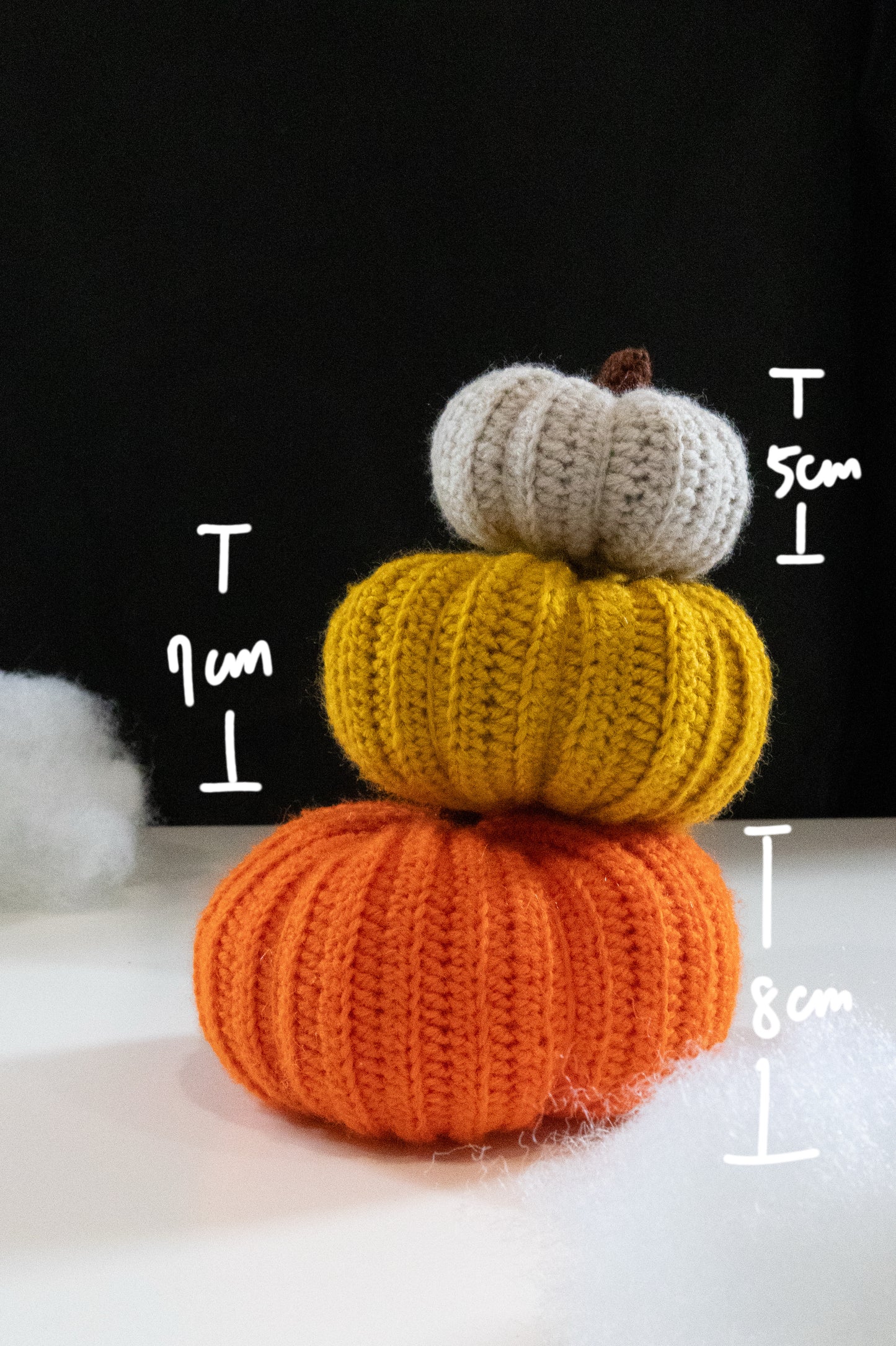 Stack of three colorful crocheted pumpkins in orange, yellow, and white, with height measurements displayed.