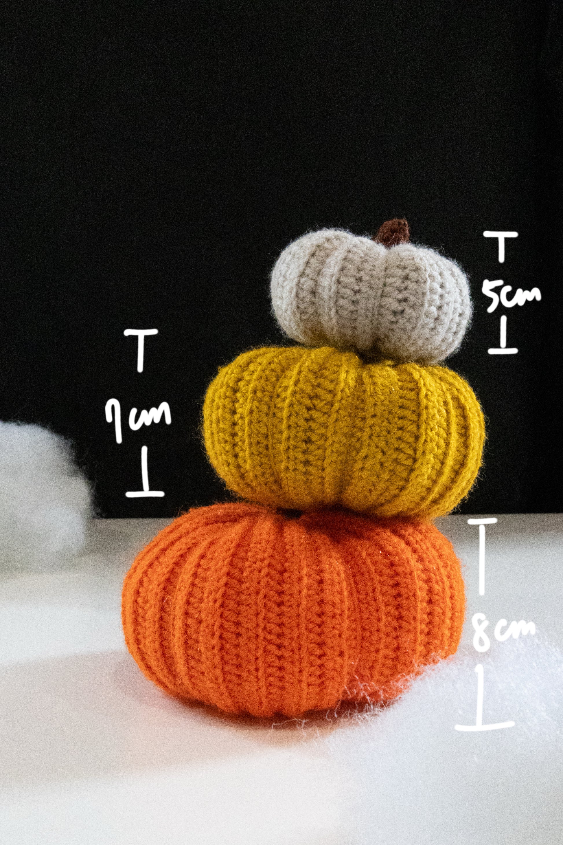 Stack of three colorful crocheted pumpkins in orange, yellow, and white, with height measurements displayed.