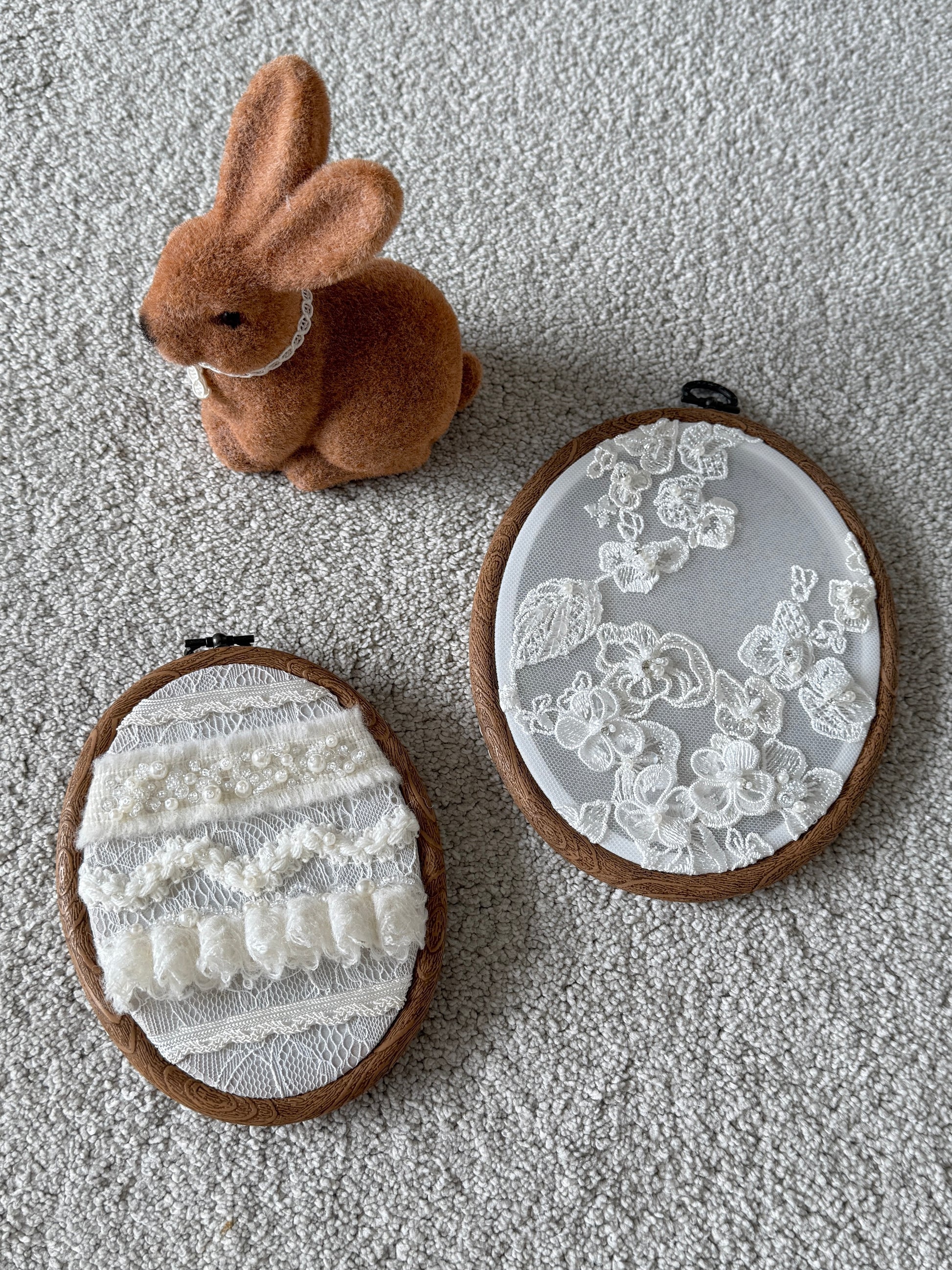 Two embroidered hoop art pieces with lace designs on a textured surface, accompanied by a small plush rabbit toy.