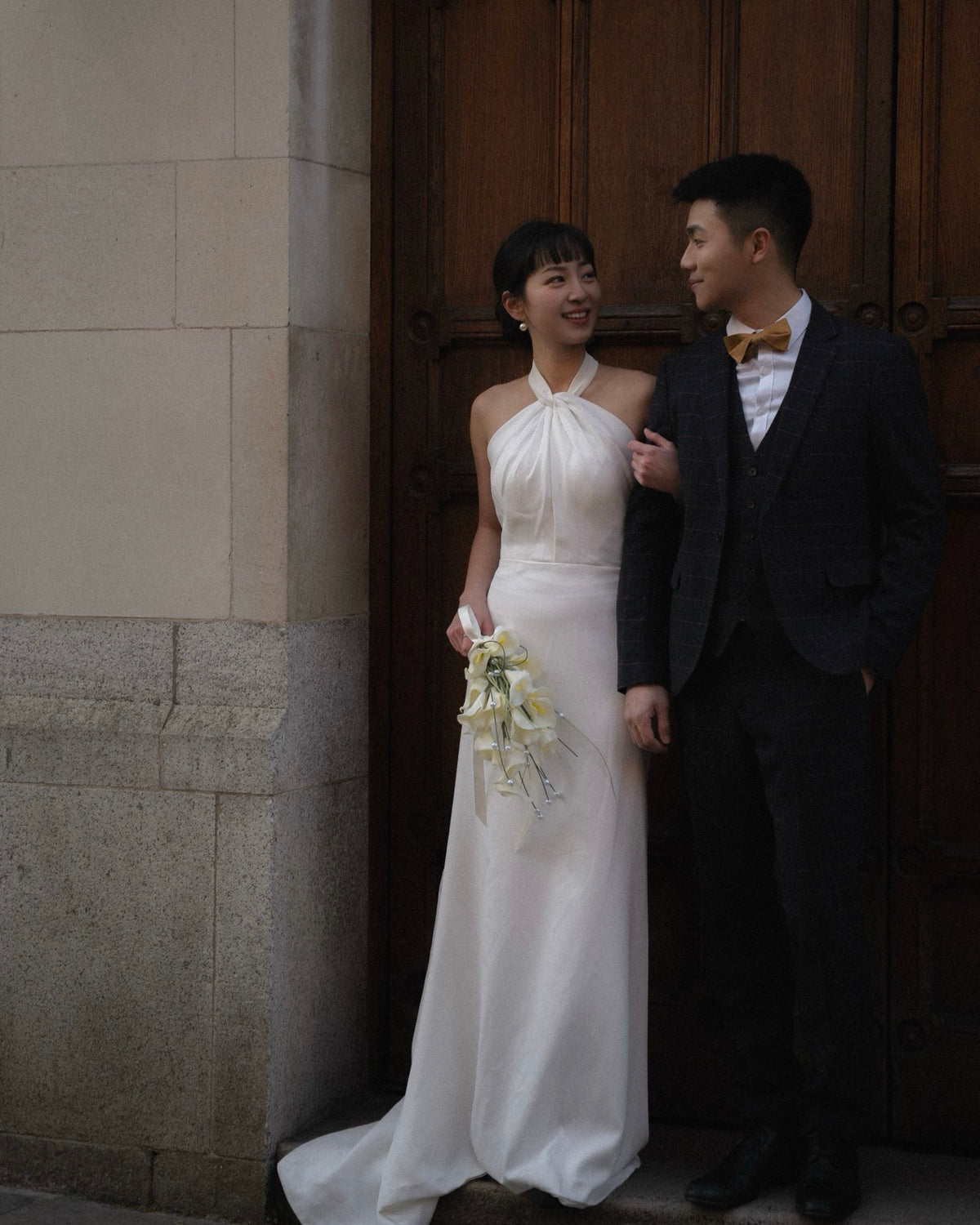 A bride in a sleeveless white halter wedding gown holding a bouquet of white flowers stands next to a groom in a dark gray suit and a yellow bow tie, posing in front of a large dark wooden door.