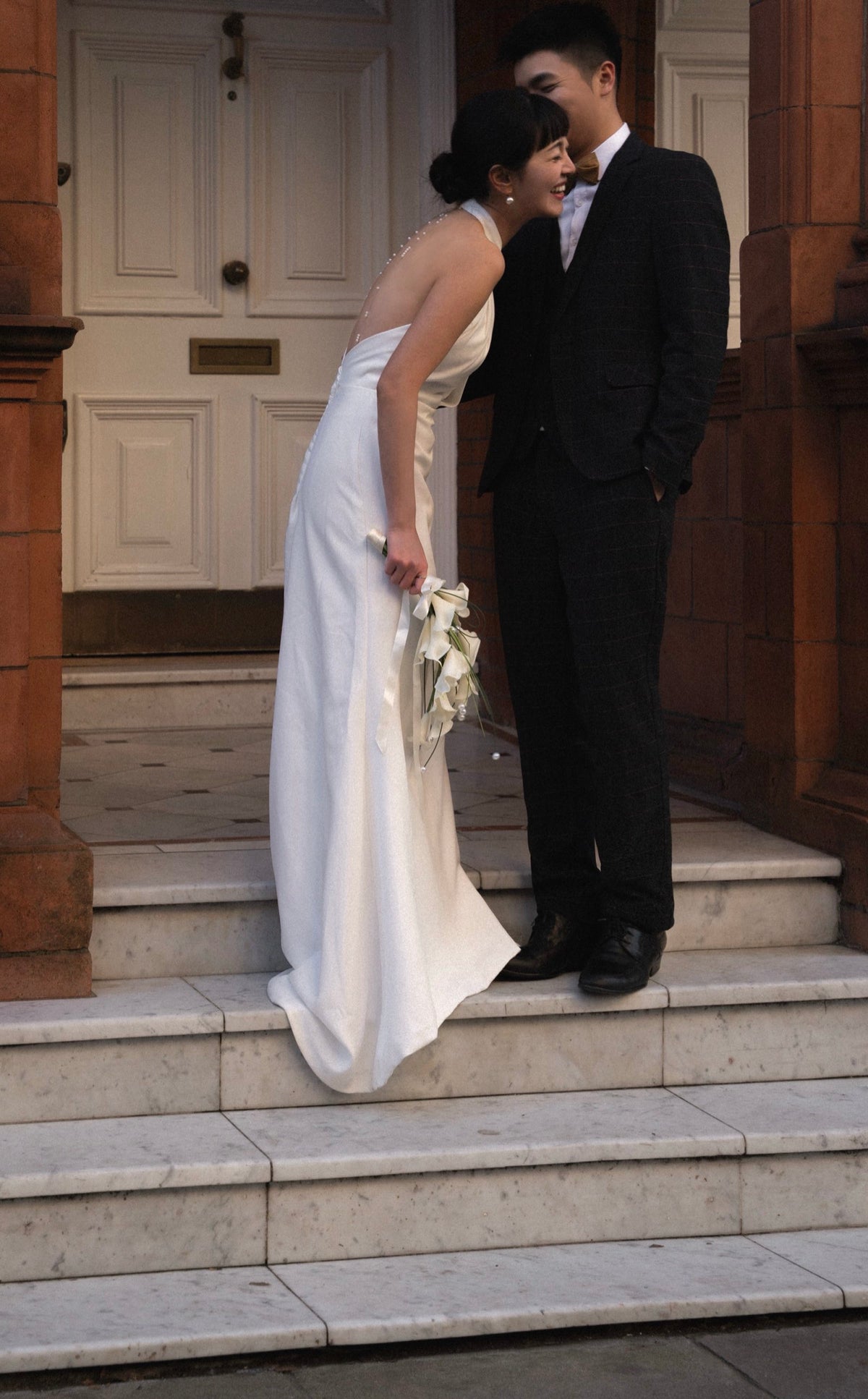 A bride in a sleeveless white wedding gown holding a bouquet of white flowers stands on stone steps beside a groom in a black suit and white shirt, posing closely in front of a building entrance.