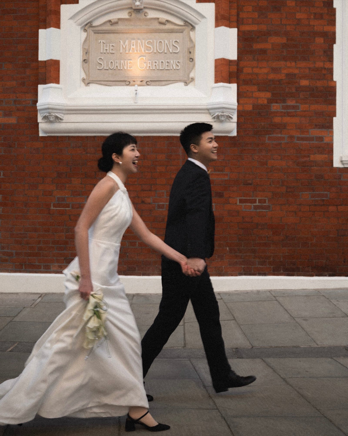 A bride in a white sleeveless wedding gown and a groom in a black suit walk hand in hand along a sidewalk in front of a red brick building with a sign reading 'The Mansions, Sloane Gardens'.