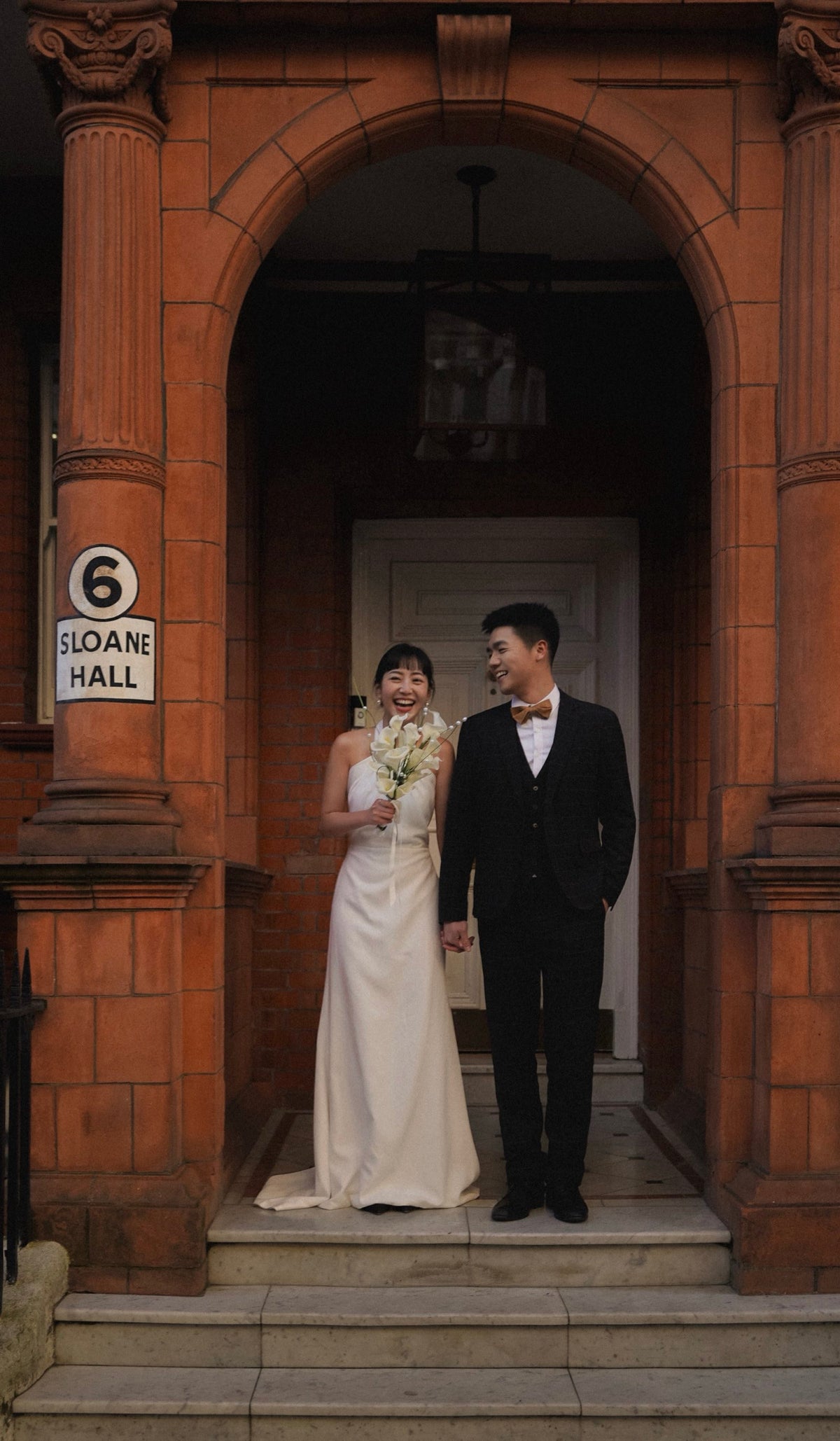 A bride in a sleeveless ivory wedding gown and a groom in a black tuxedo stand hand in hand on the steps of a red brick building with an arched doorway. A white sign on the left reads '6 SLOANE HALL'.
