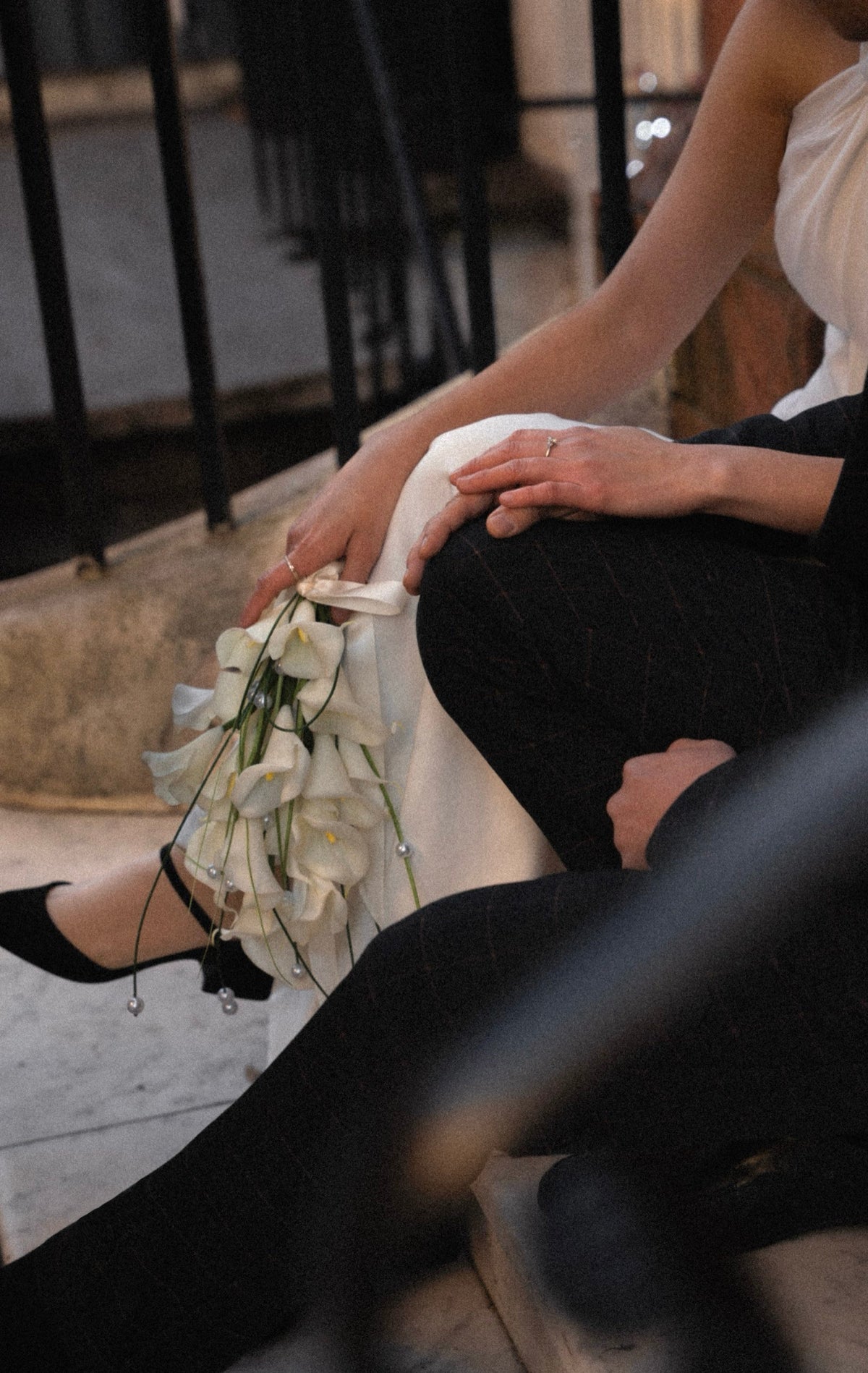 A close-up lifestyle shot of a bride sitting on stone steps, holding a bouquet of white lilies. She is wearing a white wedding dress, black pinstriped tights, and black wedding shoes.