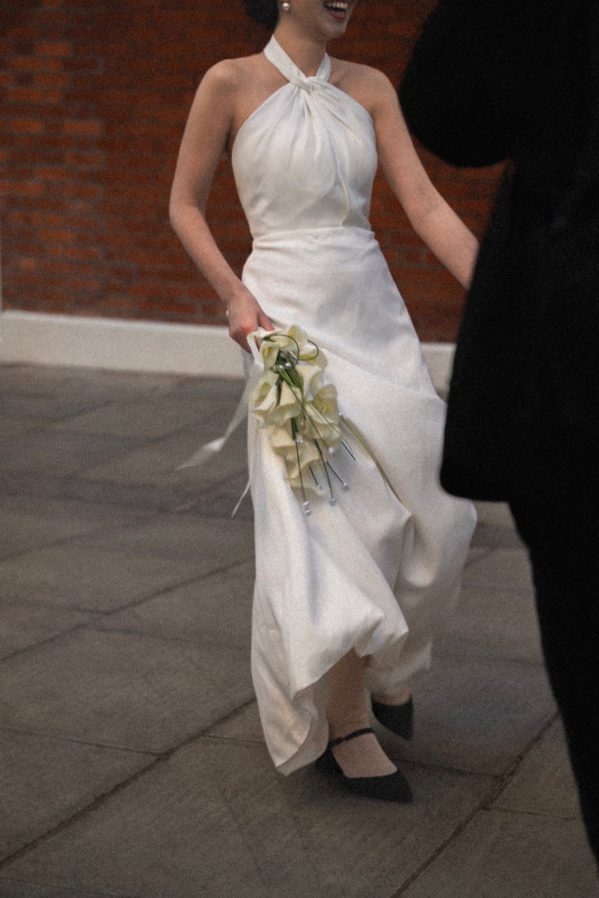A woman wearing a white halter-neck wedding dress with a gathered waist and flowing skirt, holding a bouquet of pale yellow flowers while walking on a stone pavement with a brick wall in the background.