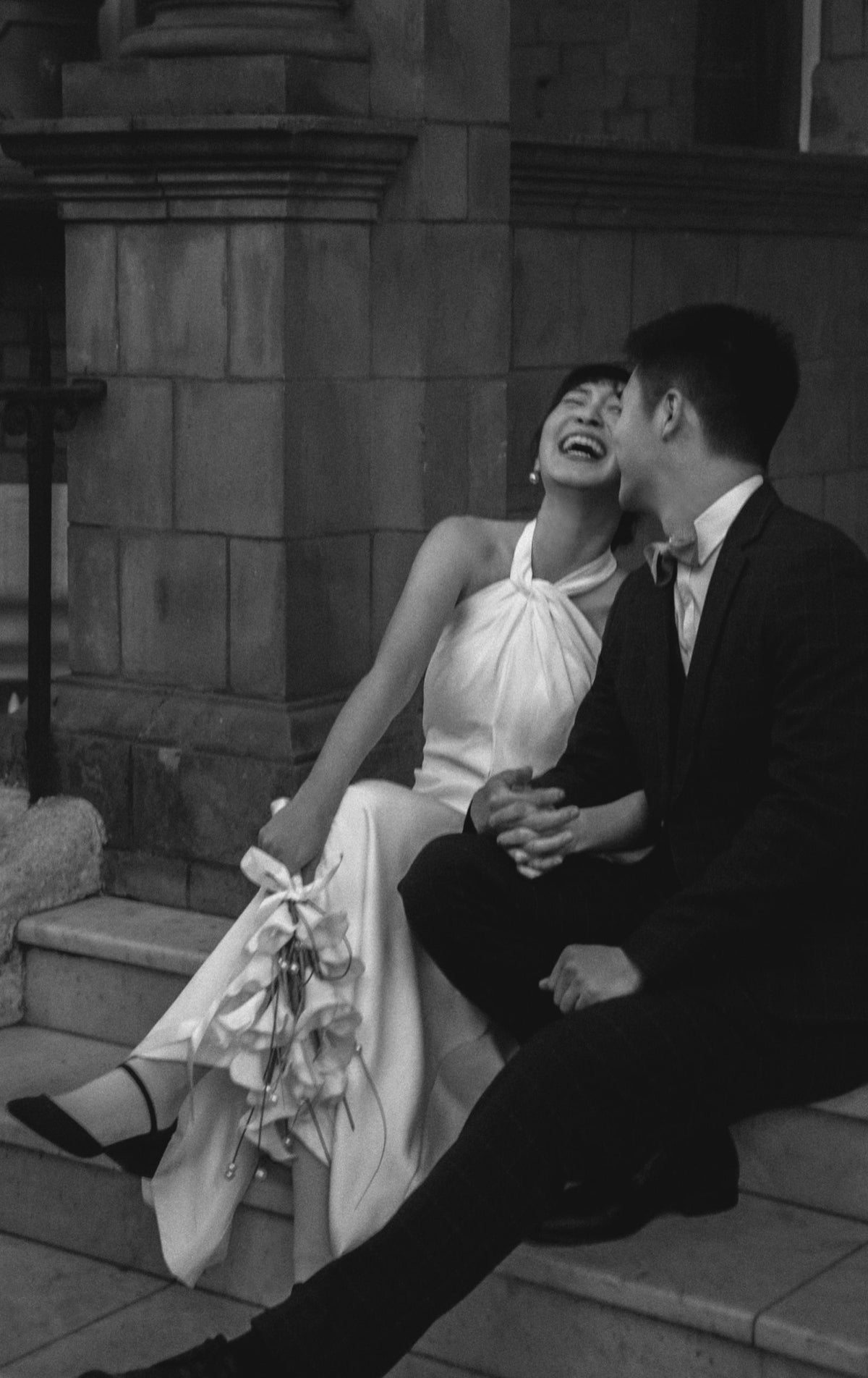 Black and white wedding photograph of a smiling bride in a sleeveless halter gown sitting on stone steps beside a groom in a dark suit.