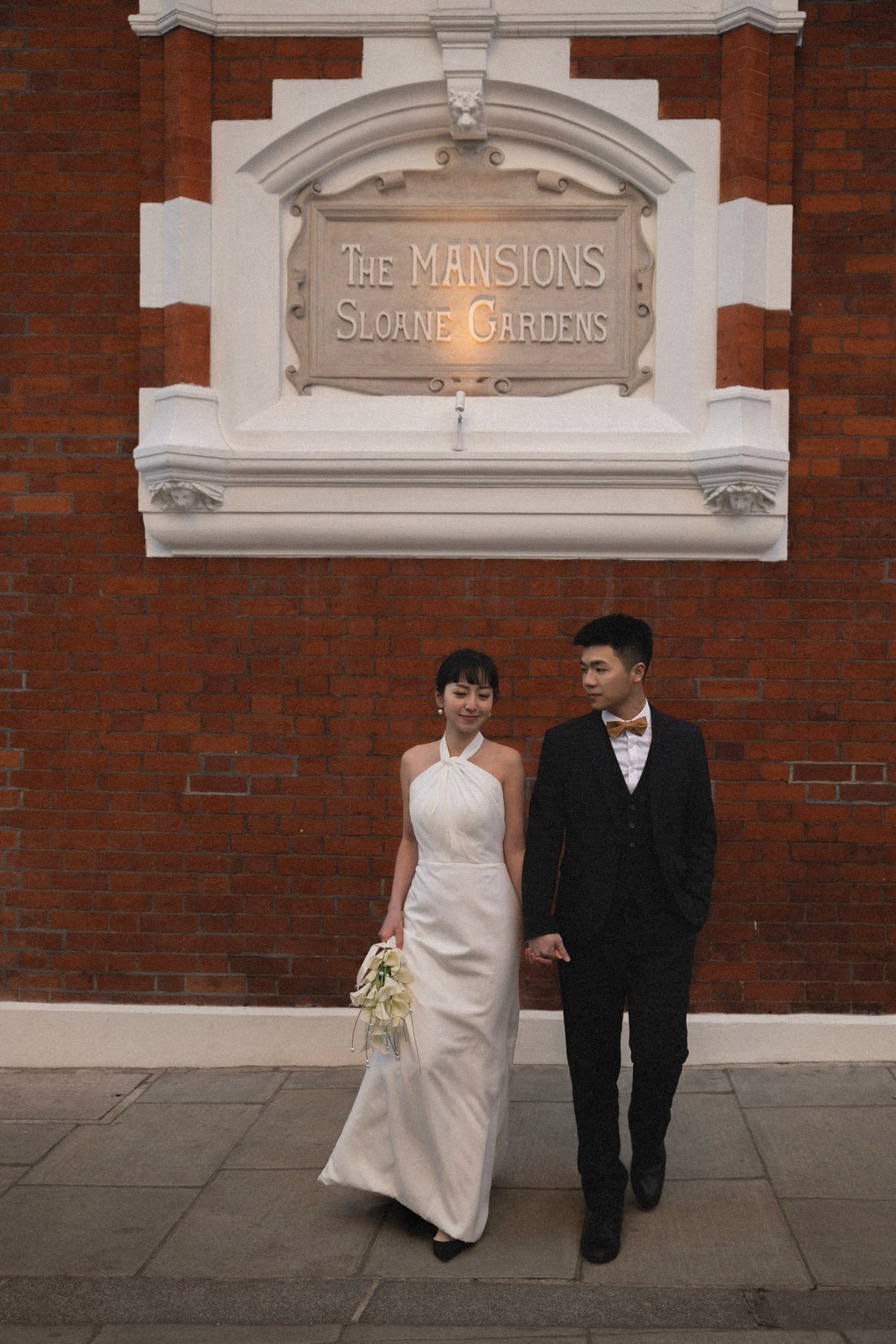 A bride in a sleeveless white wedding gown and a groom in a black suit walk hand in hand in front of a red brick building with a white sign reading 'The Mansion's Sloane Gardens'.