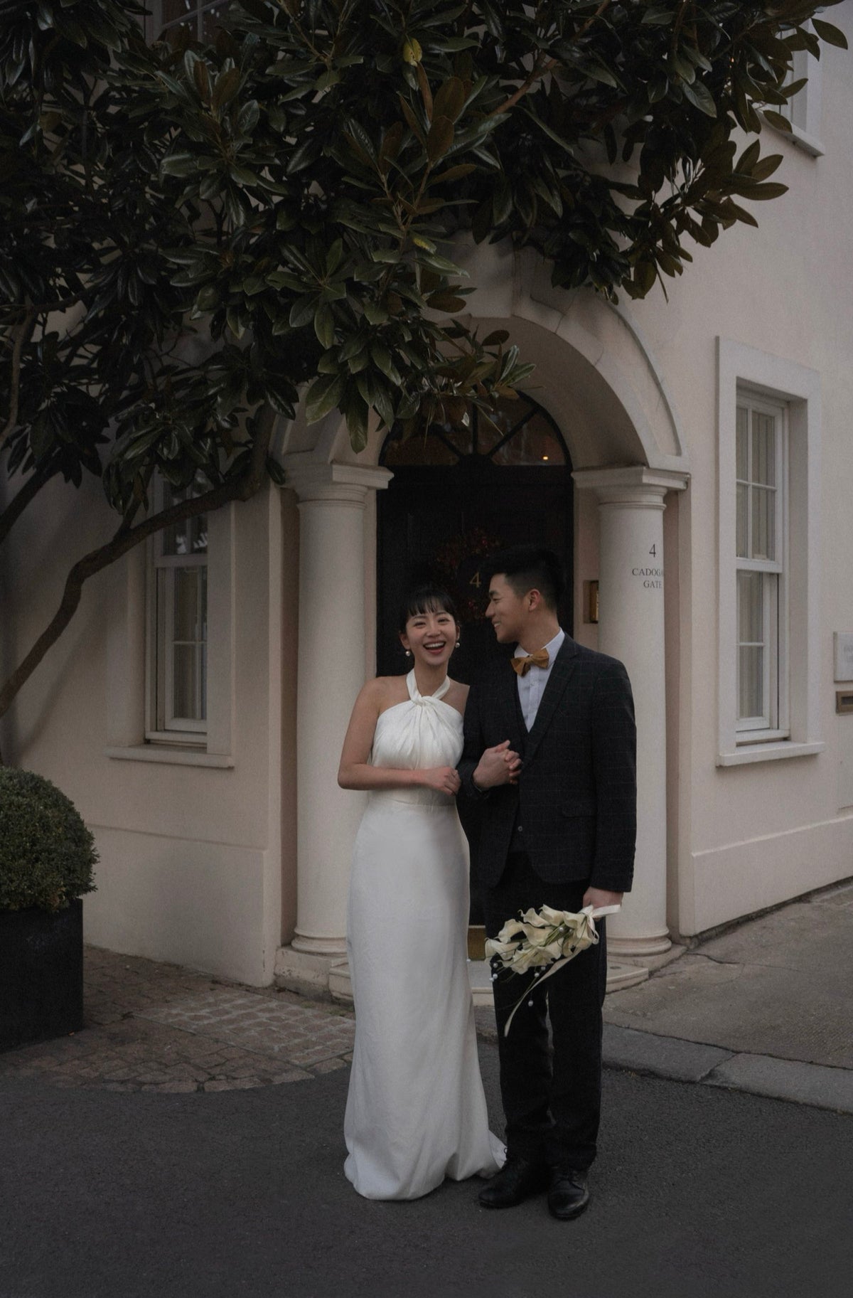 A bride in a sleeveless white wedding gown and a groom in a black suit with a yellow bow tie stand together in front of a cream-colored building with an arched doorway and leafy tree branches overhead.