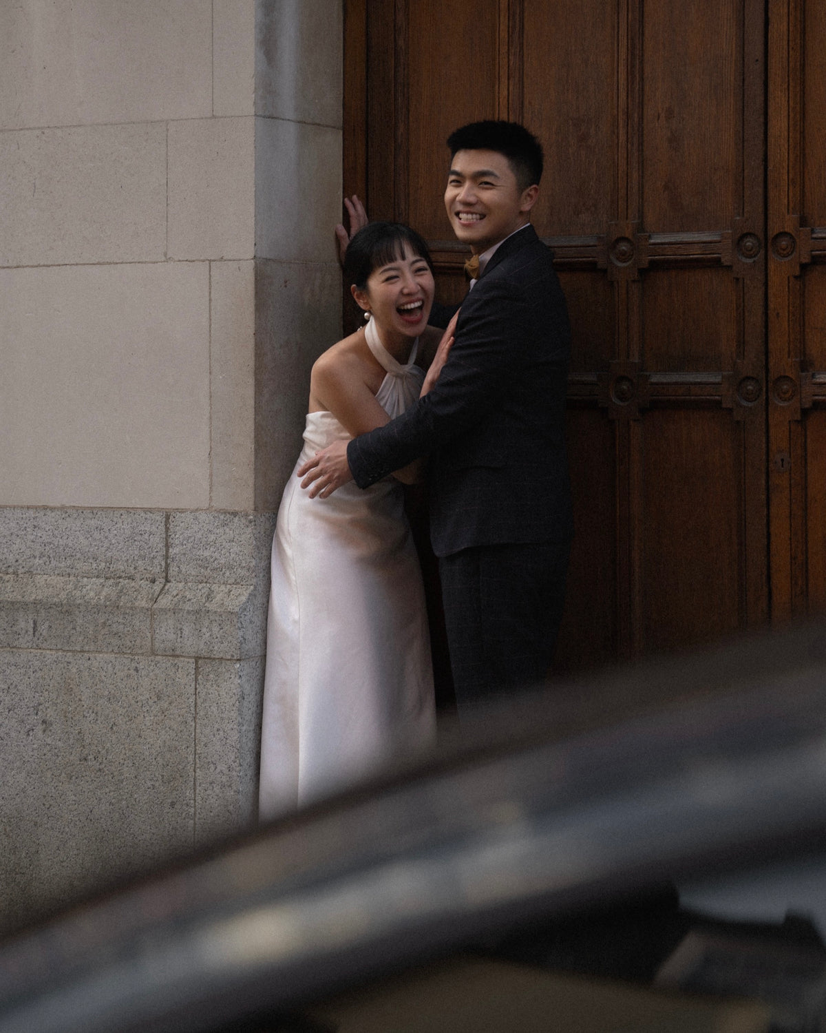 A smiling couple embracing in front of a large wooden door. The woman wears a sleeveless white wedding gown, and the man wears a dark suit.