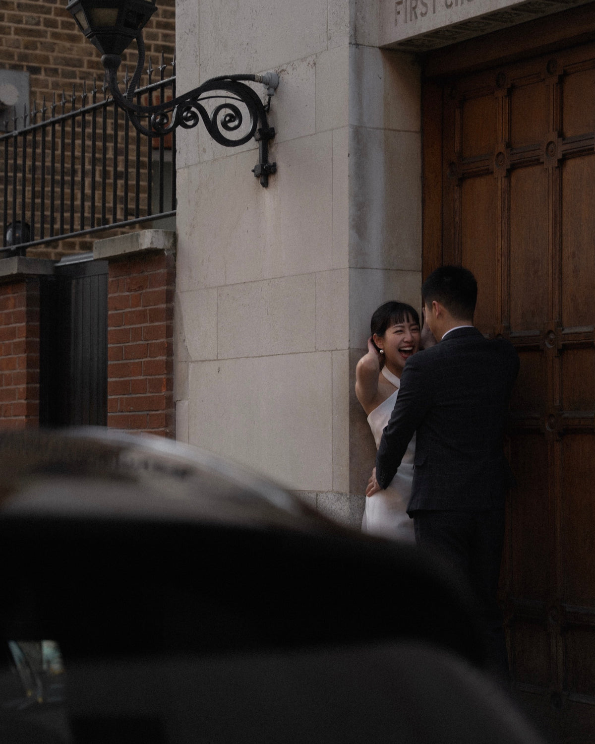 A bride in a white wedding dress and a groom in a dark suit embrace and laugh beside a large wooden door and stone pillar.