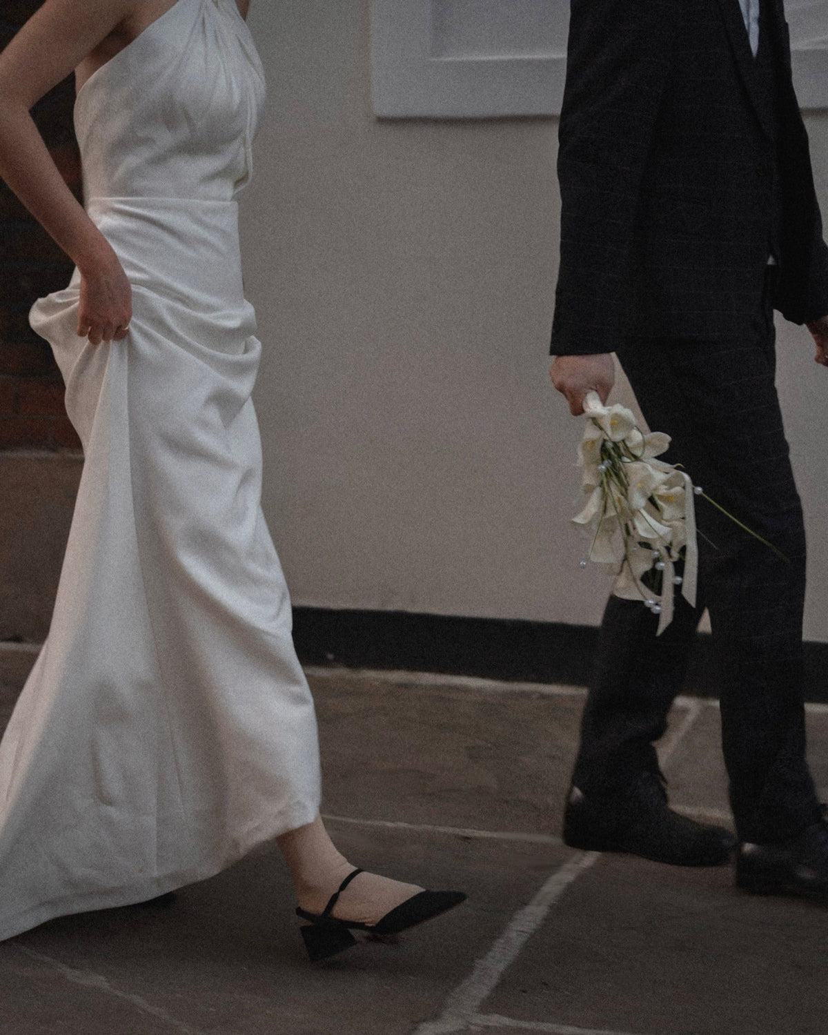 A woman in a draped white wedding dress walking outdoors, holding a white bouquet with a man in a dark suit beside her.