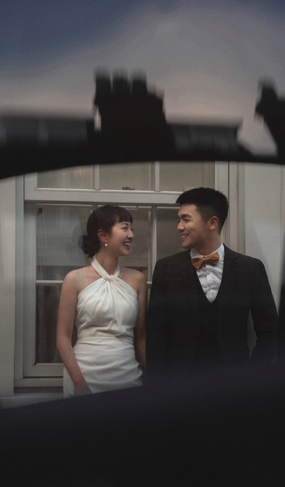 A couple smiling at each other in front of a window, framed by a dark silhouette at the top and bottom. The woman wears a white halter-neck dress, and the man wears a dark suit with a brown bow tie.