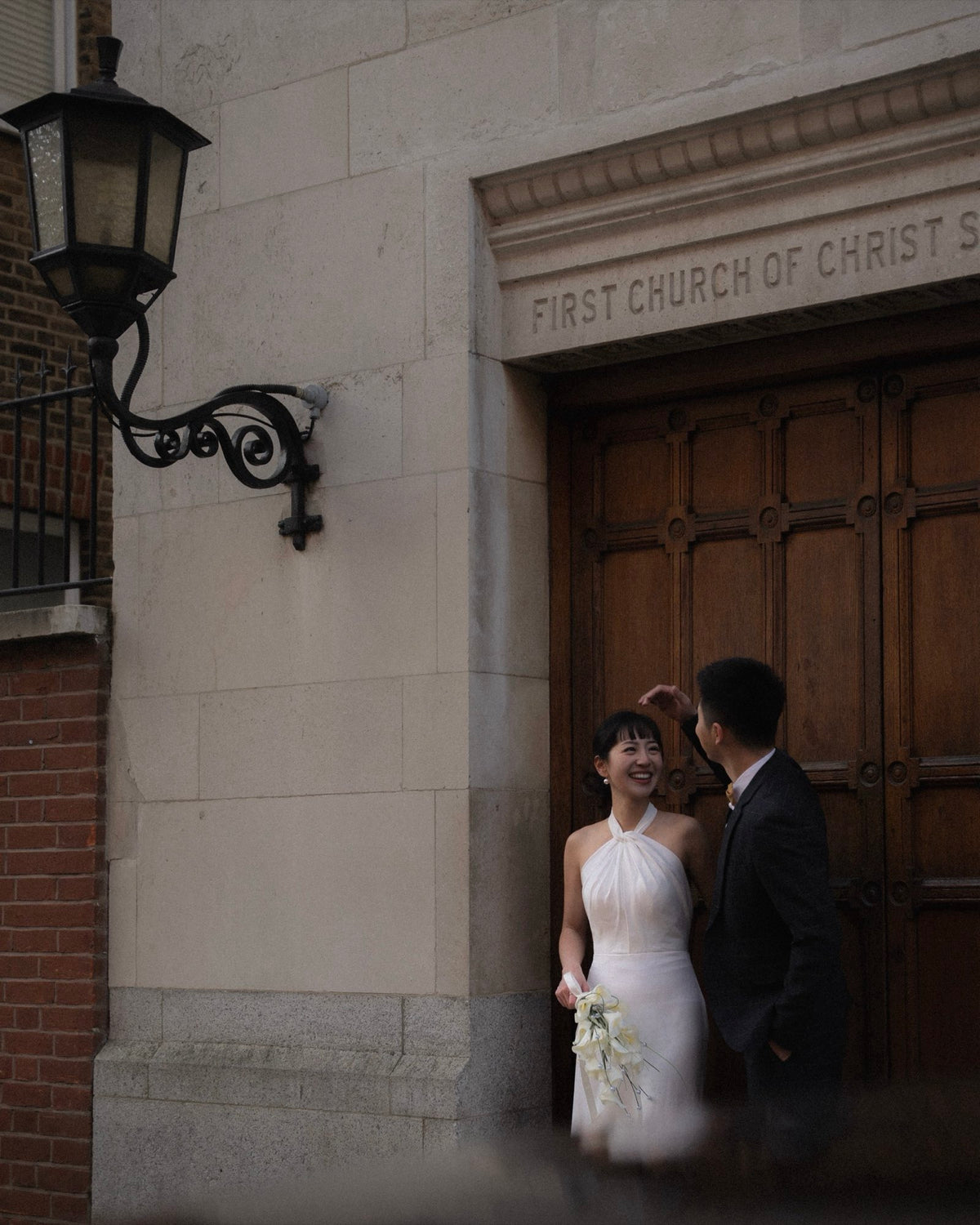 A bride in a white halter-neck wedding gown and a groom in a dark suit pose in front of the wooden double doors of the First Church of Christ Saviour, with a stone wall and a black wall lamp on the left.