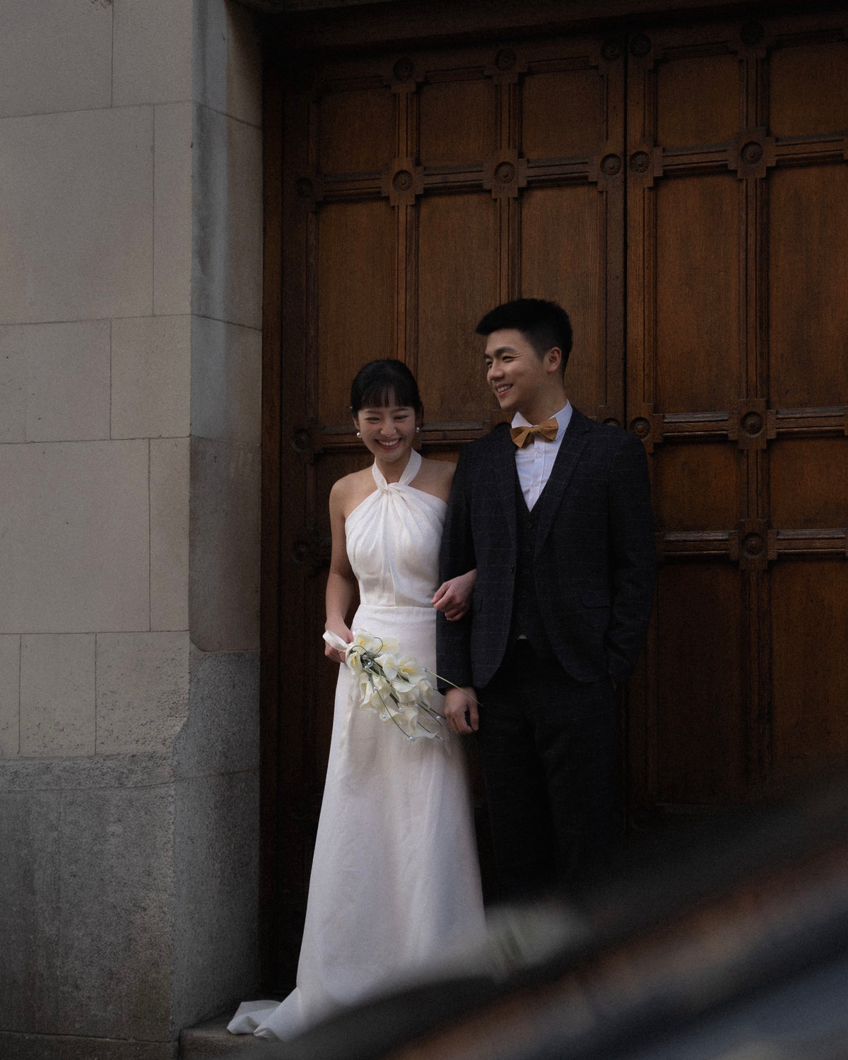 A bride in a white halter-neck wedding gown holding a bouquet of white flowers stands beside a groom in a dark suit and bow tie in front of a large wooden door.
