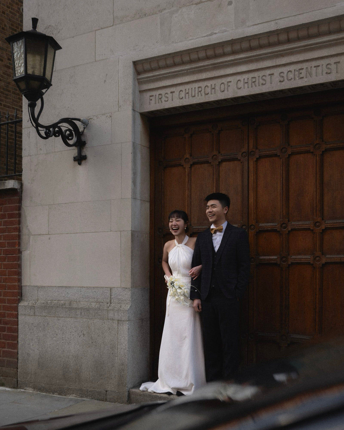 A bride in a sleeveless white wedding gown holding a bouquet of white flowers stands beside a groom in a dark suit with a bow tie, posing in front of a large wooden door of the First Church of Christ Scientist.