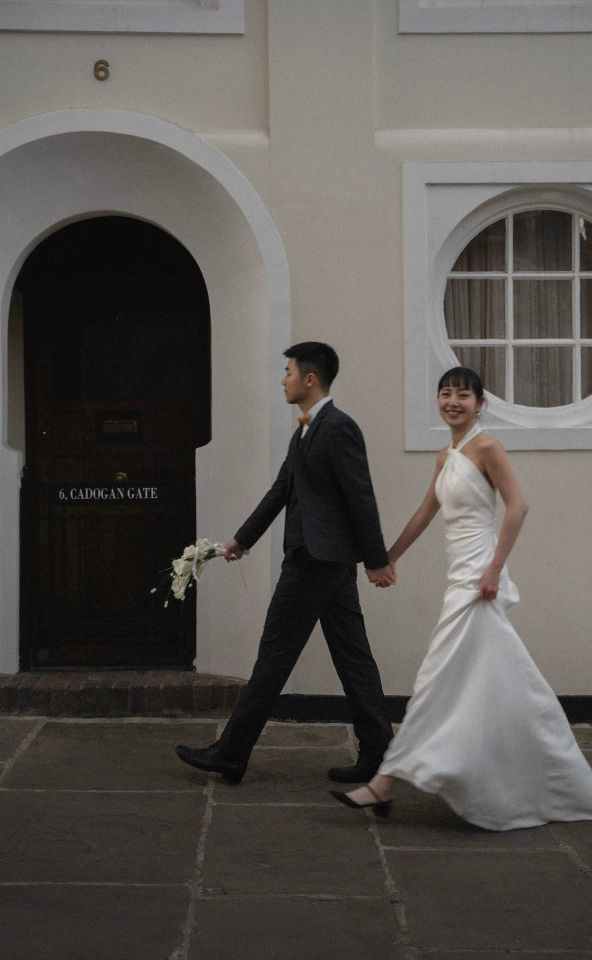 A bride in a white wedding gown and a groom in a dark suit walk hand in hand outside a building with white arched doorways and a dark wooden door labeled 'A. CADOGAN GATE'.