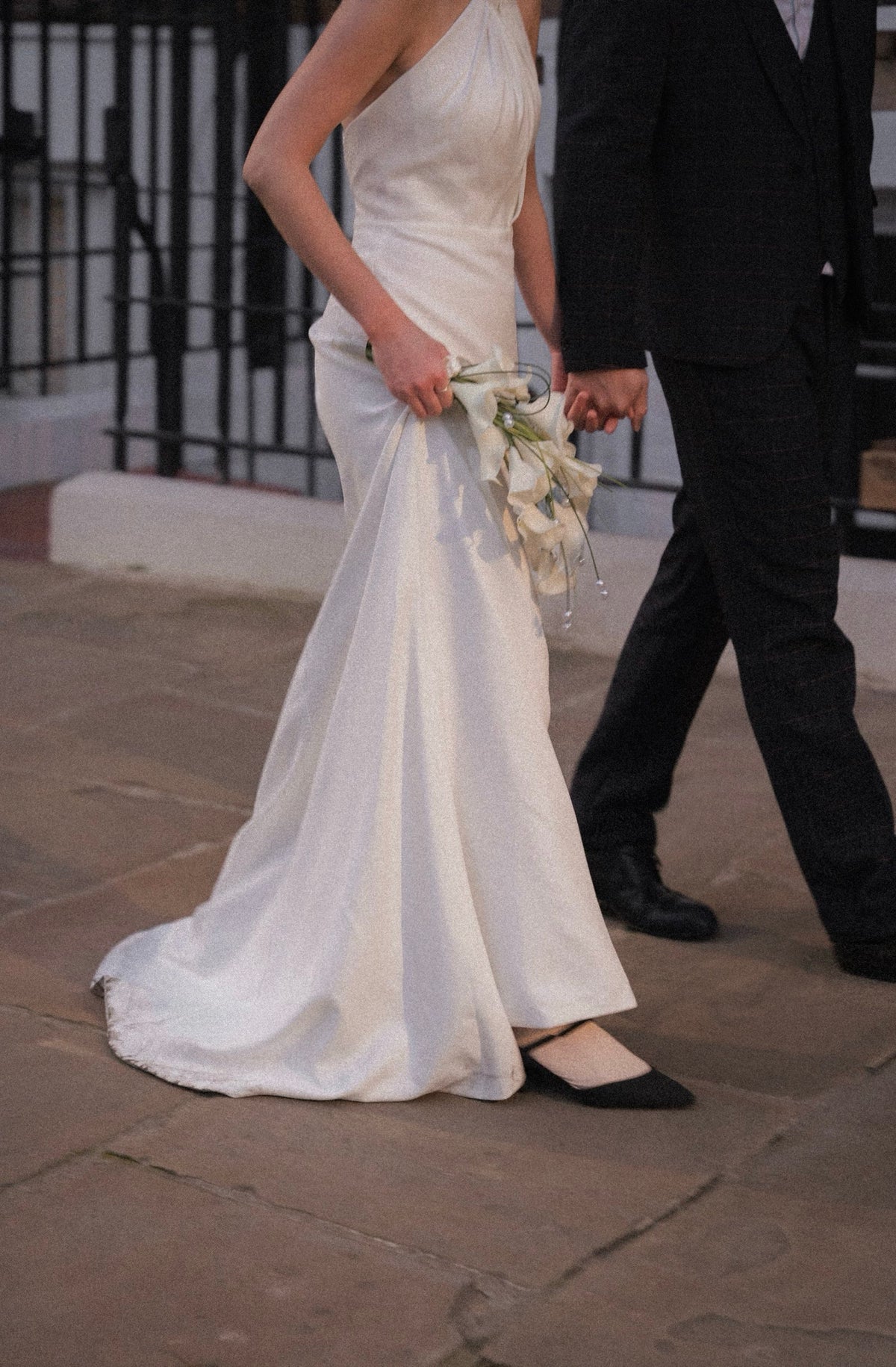 A woman in a sleeveless white wedding gown holding a bouquet of white flowers, walking outdoors and holding hands with a man in a dark suit. The photo is cropped from the waist down, showing her black flats on a stone pavement.