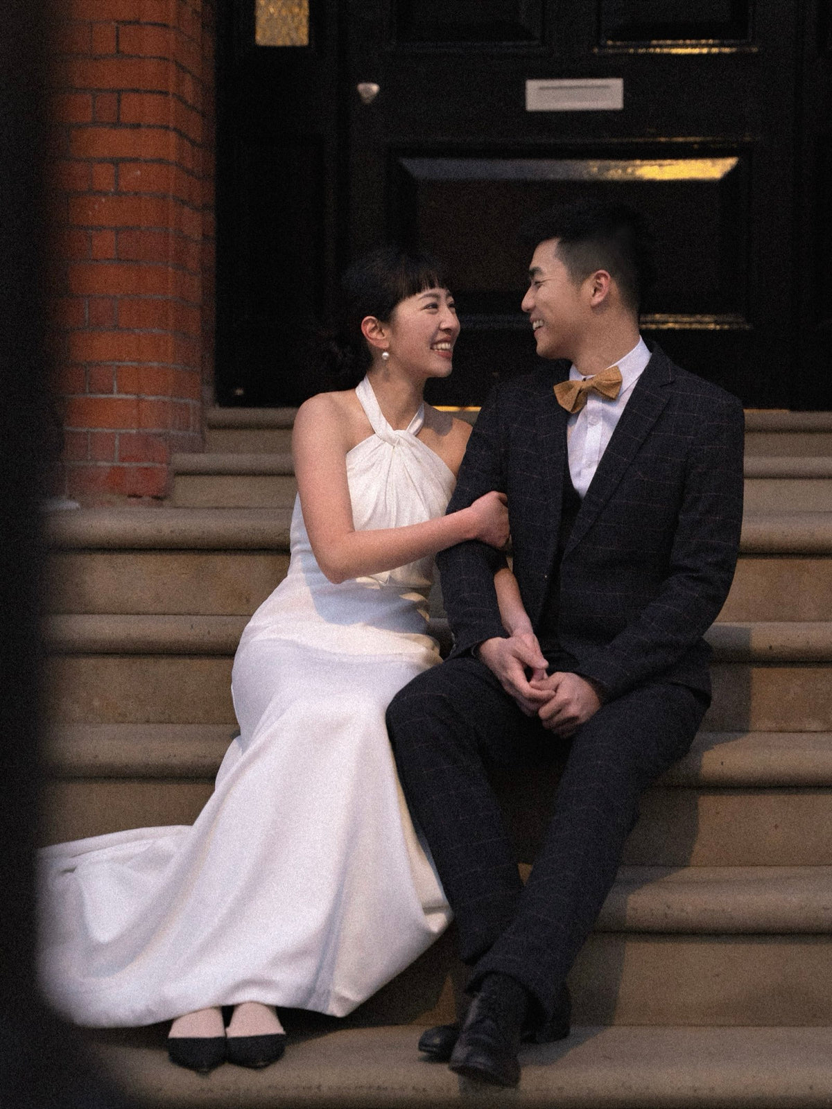 A couple sitting together on stone steps in front of a building entrance. The woman wears a sleeveless white wedding gown, and the man wears a dark textured suit with a yellow bow tie.