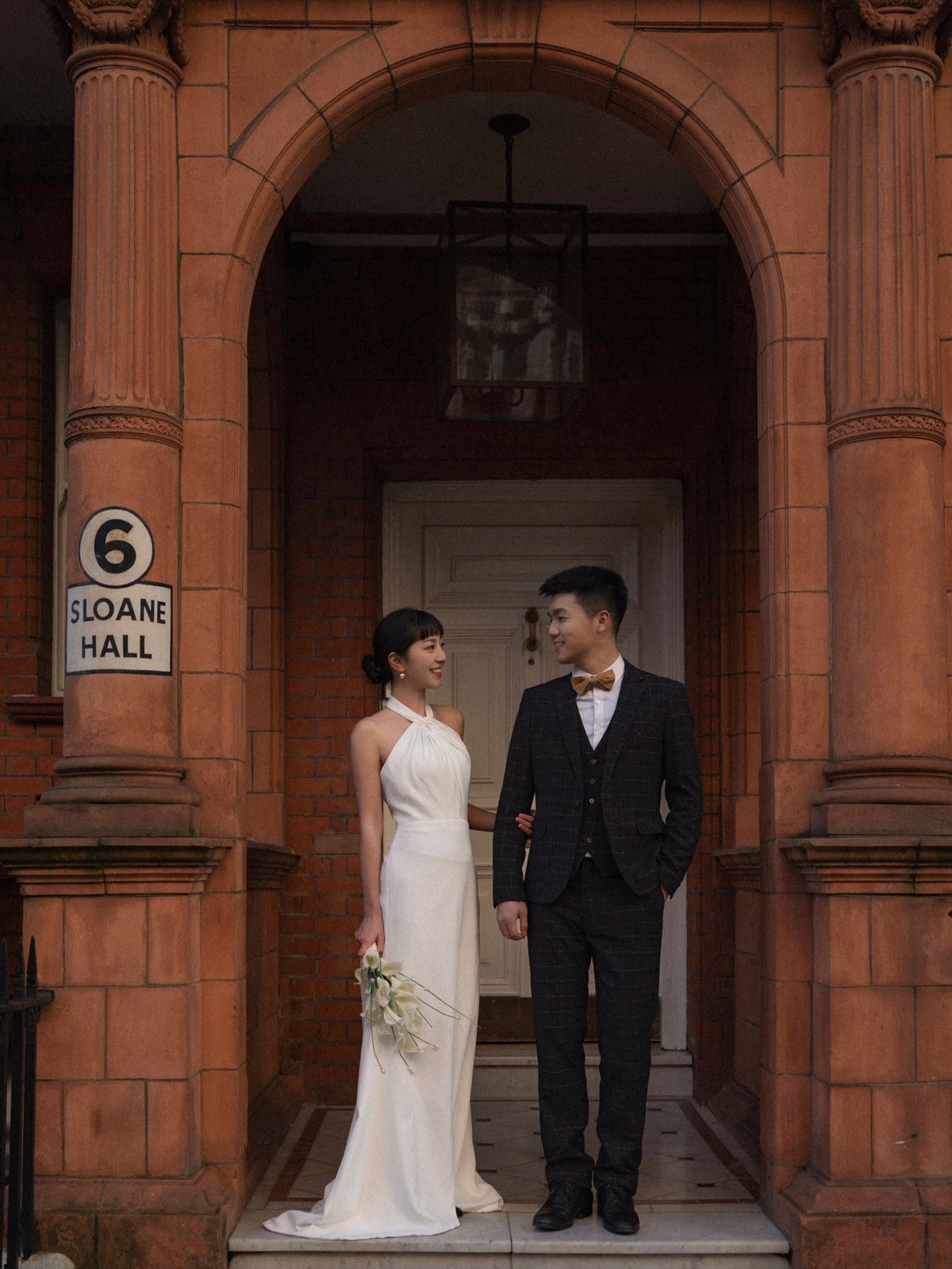 A bride in a sleeveless white wedding gown and a groom in a dark suit stand together on the steps of a red brick building entrance with an arched doorway. A sign on the left reads '6 SLOANE HALL'.