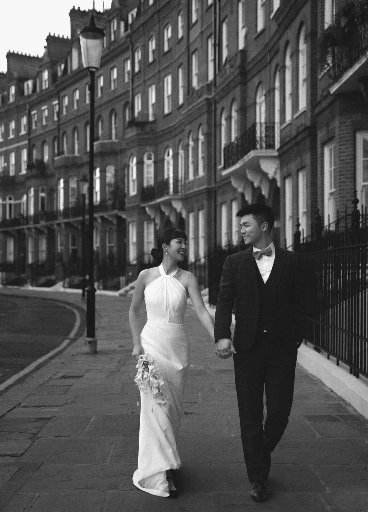 A black and white photograph of a bride in a sleeveless white wedding gown and a groom in a dark suit walking hand-in-hand down a London street lined with classic apartment buildings.