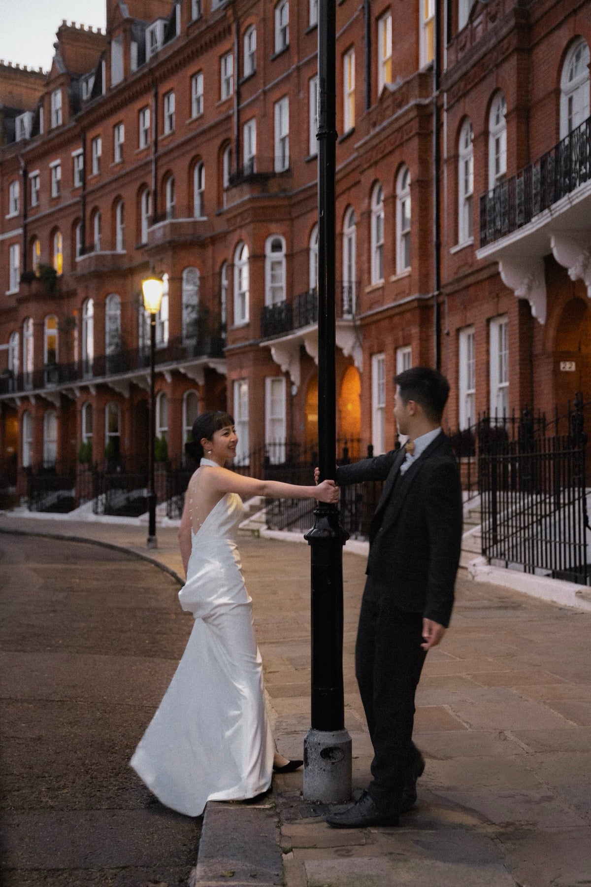 A bride in a white sleeveless wedding gown and a groom in a dark suit stand on a city street in front of red brick apartment buildings, holding hands beside a black lamppost.