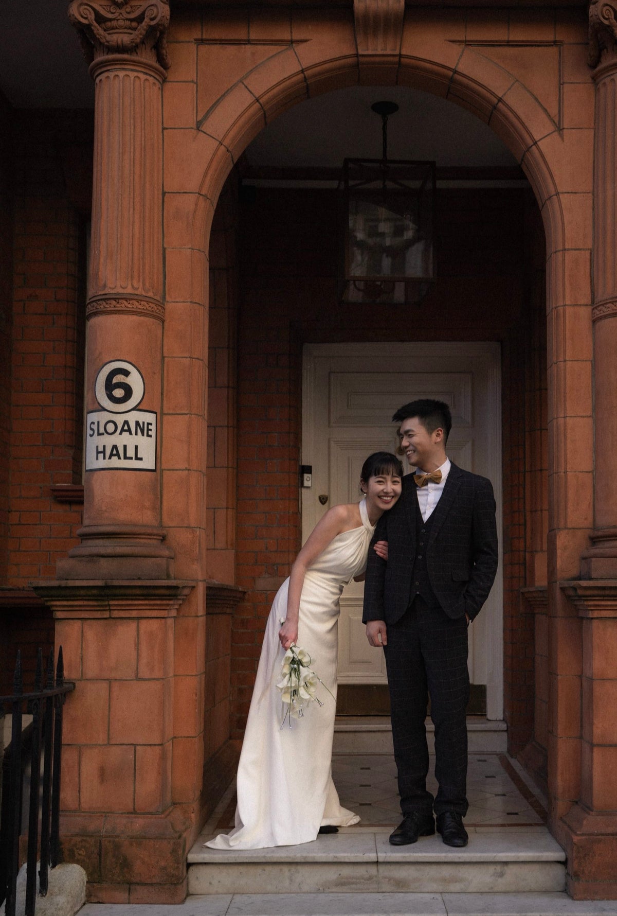 A bride in a sleeveless ivory wedding gown and a groom in a black suit with a white bow tie stand together in the doorway of a red brick building. A sign on the left pillar reads '6 SLOANE HALL'.