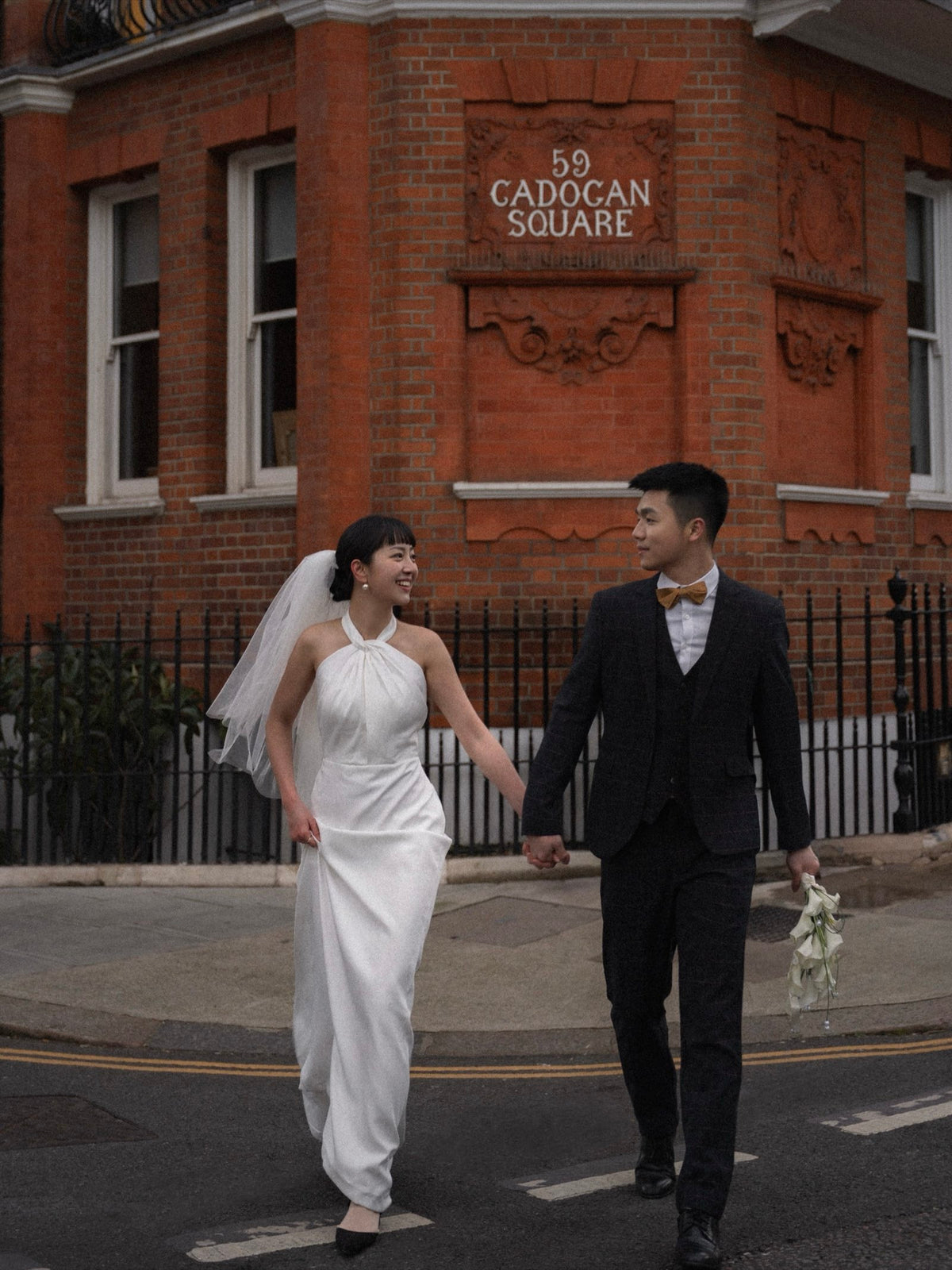A bride in a white halter-neck wedding dress with a veil and a groom in a black suit walking hand-in-hand down a street in front of a red brick building labeled '59 CADOCAN SQUARE'.