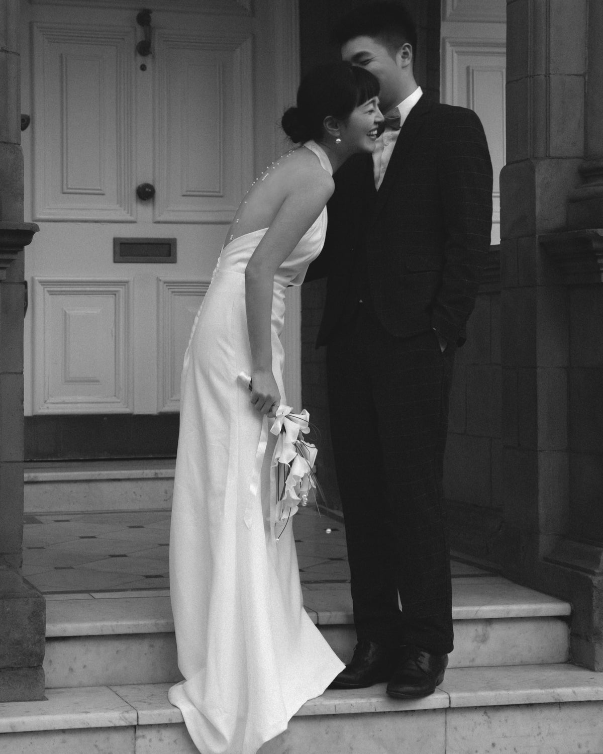 A black and white photograph of a bride in a sleeveless white wedding gown leaning affectionately against a groom in a dark suit on stone steps in front of a building door.
