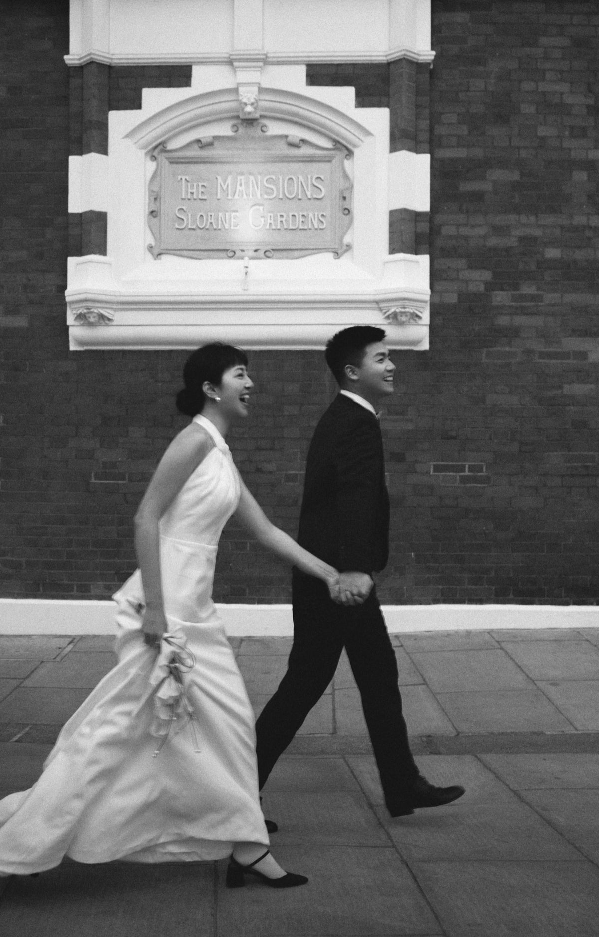 A black and white photograph of a bride and groom walking hand-in-hand outdoors in front of a brick building with a sign reading 'The Mansions Sloane Gardens'.