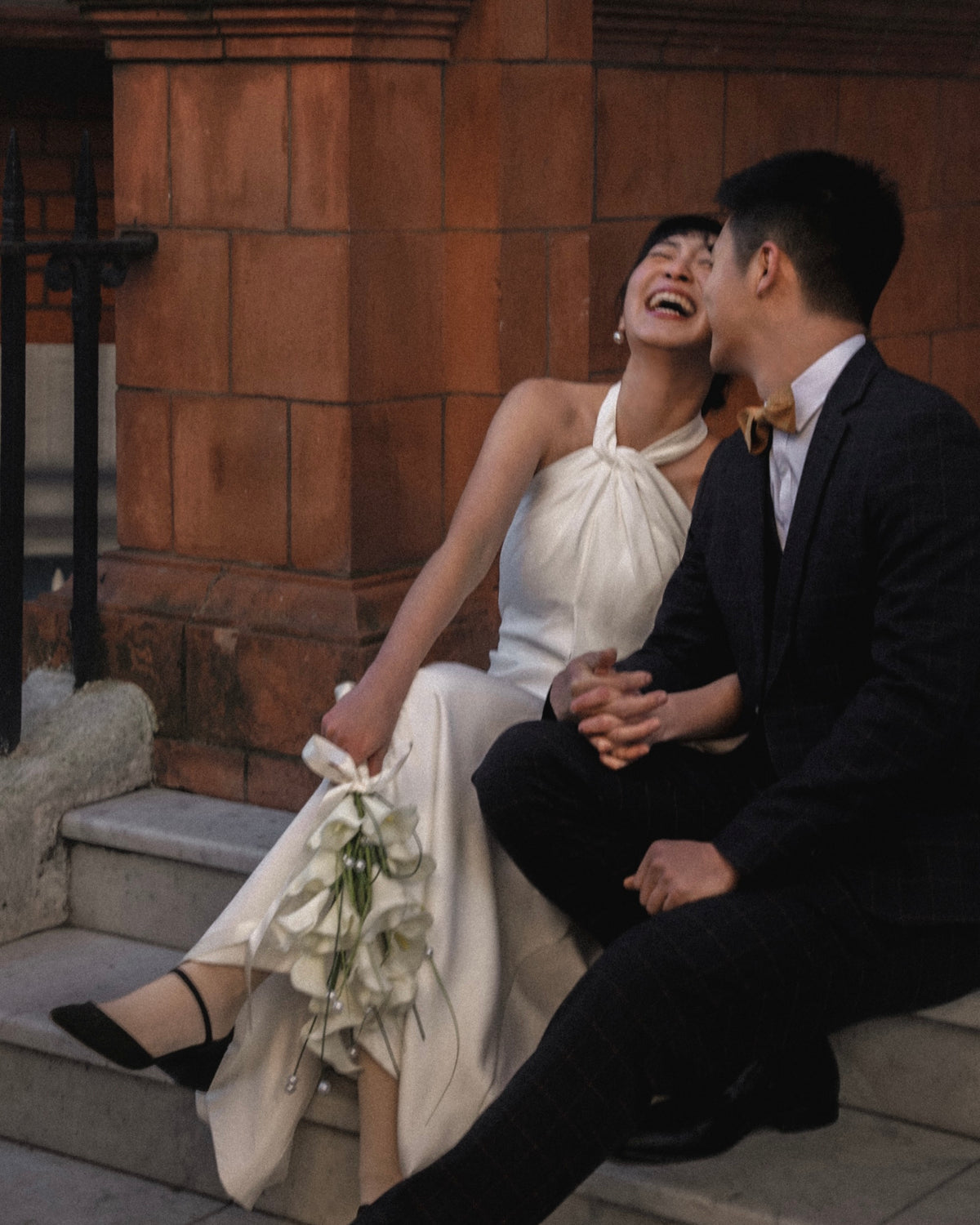 A bride in a cream halter-neck wedding dress and a groom in a dark suit sit together on stone steps, laughing and holding hands. The bride holds a bouquet of white flowers.
