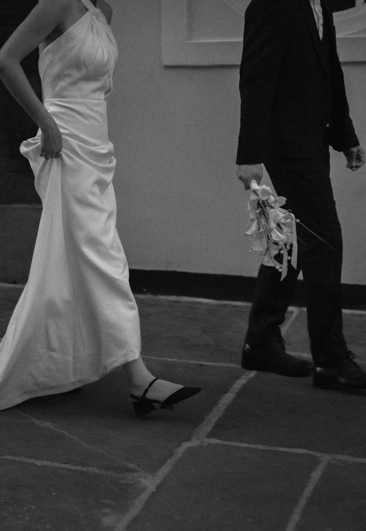 Black and white photograph of a bride in a sleeveless draped wedding gown walking beside a groom in a dark suit, holding a bouquet of flowers on a stone pavement.