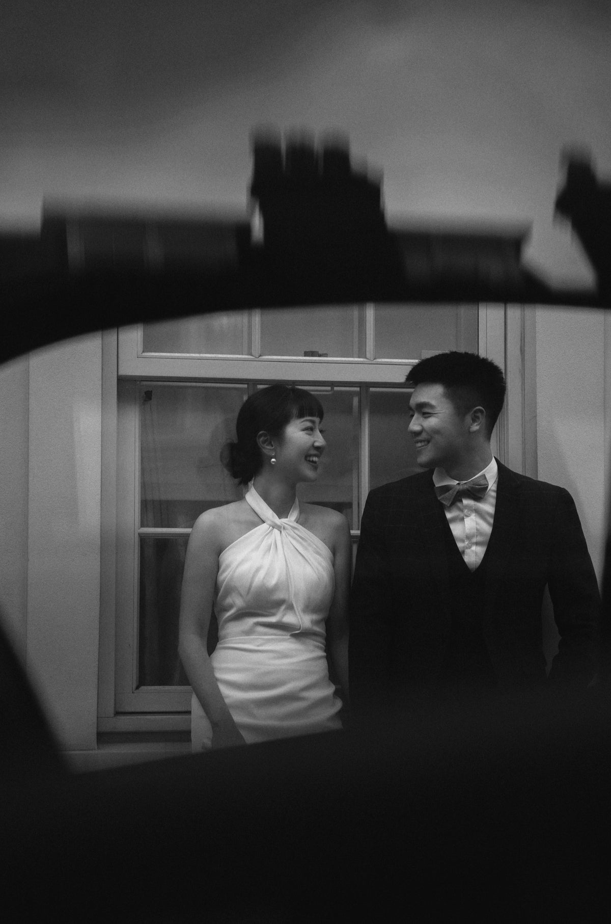 Black and white wedding photograph of a bride and groom standing side by side, framed by a dark silhouette in the foreground.