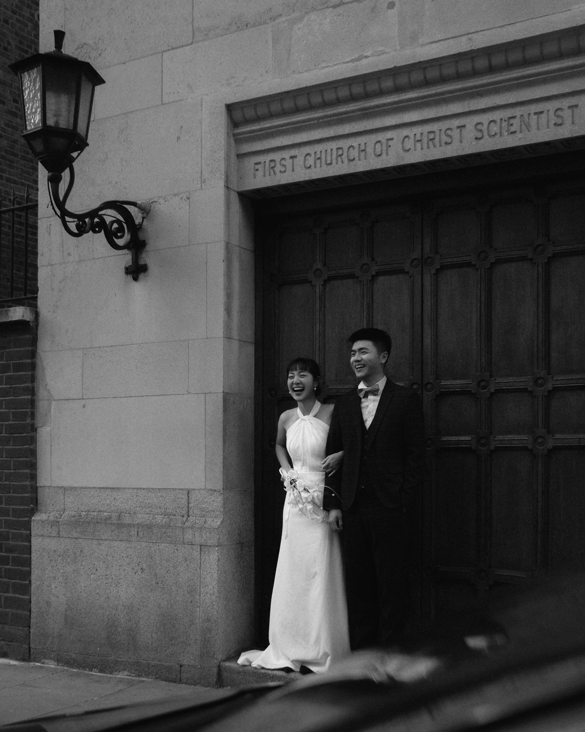 A black and white photograph of a bride in a sleeveless white wedding gown and a groom in a dark suit standing together in front of a large wooden church door with the inscription 'FIRST CHURCH OF CHRIST SCIENTIS'.