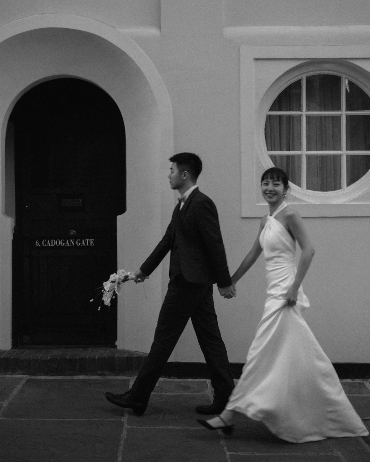 A black and white photograph of a bride in a sleeveless white wedding gown and a groom in a dark suit walking hand-in-hand outdoors in front of a building with an arched doorway and a round window.
