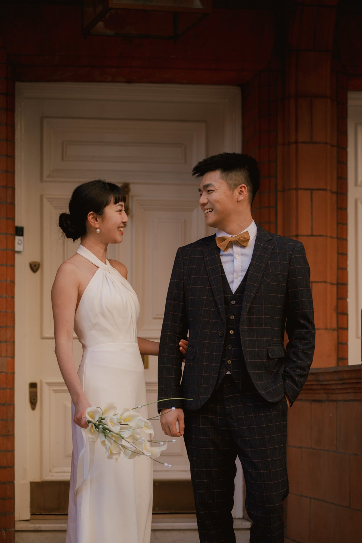 A bride in a white halter-neck wedding dress holding a bouquet of white lilies stands next to a groom in a dark plaid three-piece suit with a yellow bow tie, posing in front of a brick and wood door.