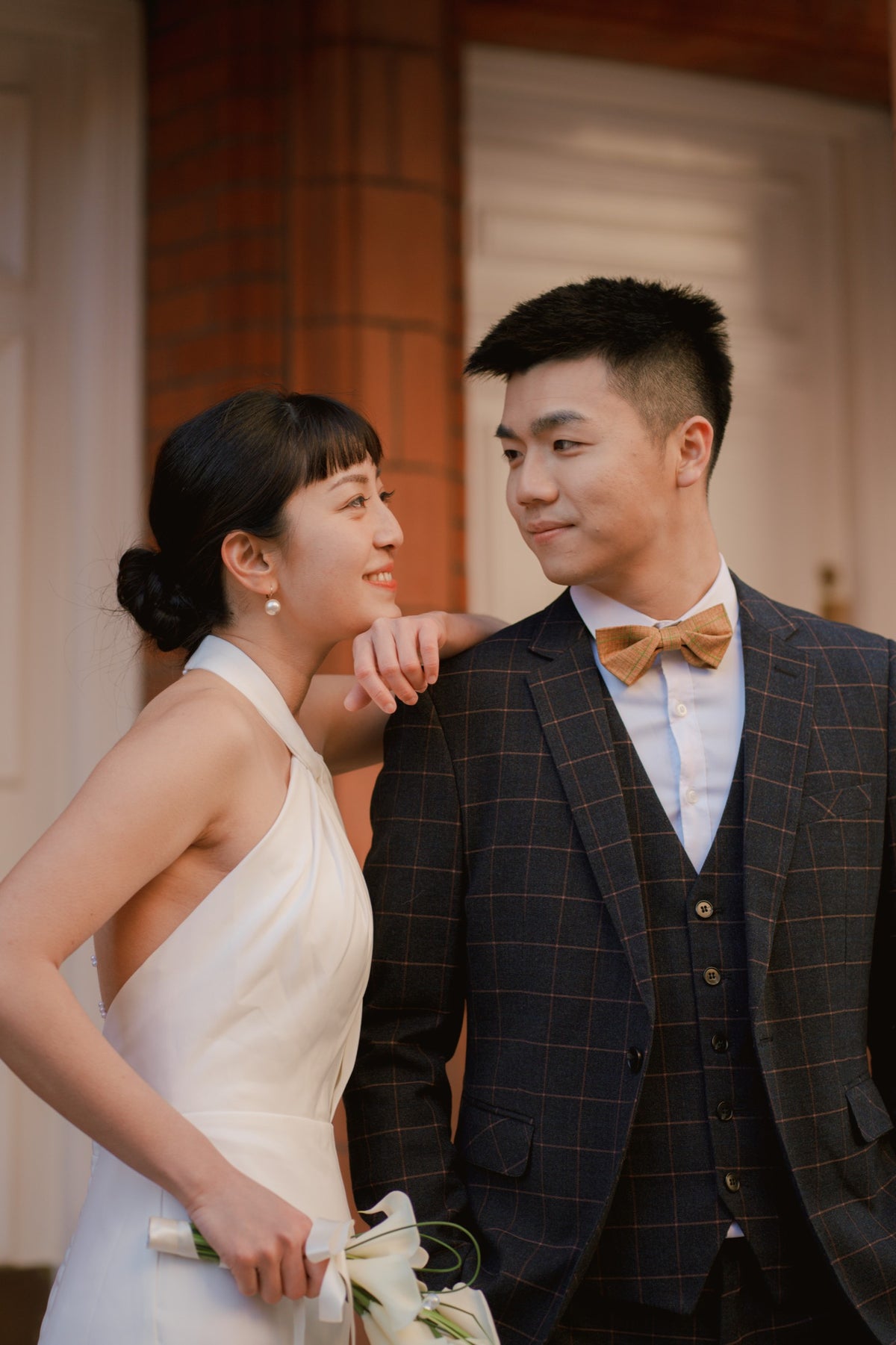A bride in a white halter-neck wedding dress and a groom in a dark plaid suit with a tan bow tie pose together in front of a warm-toned building doorway.