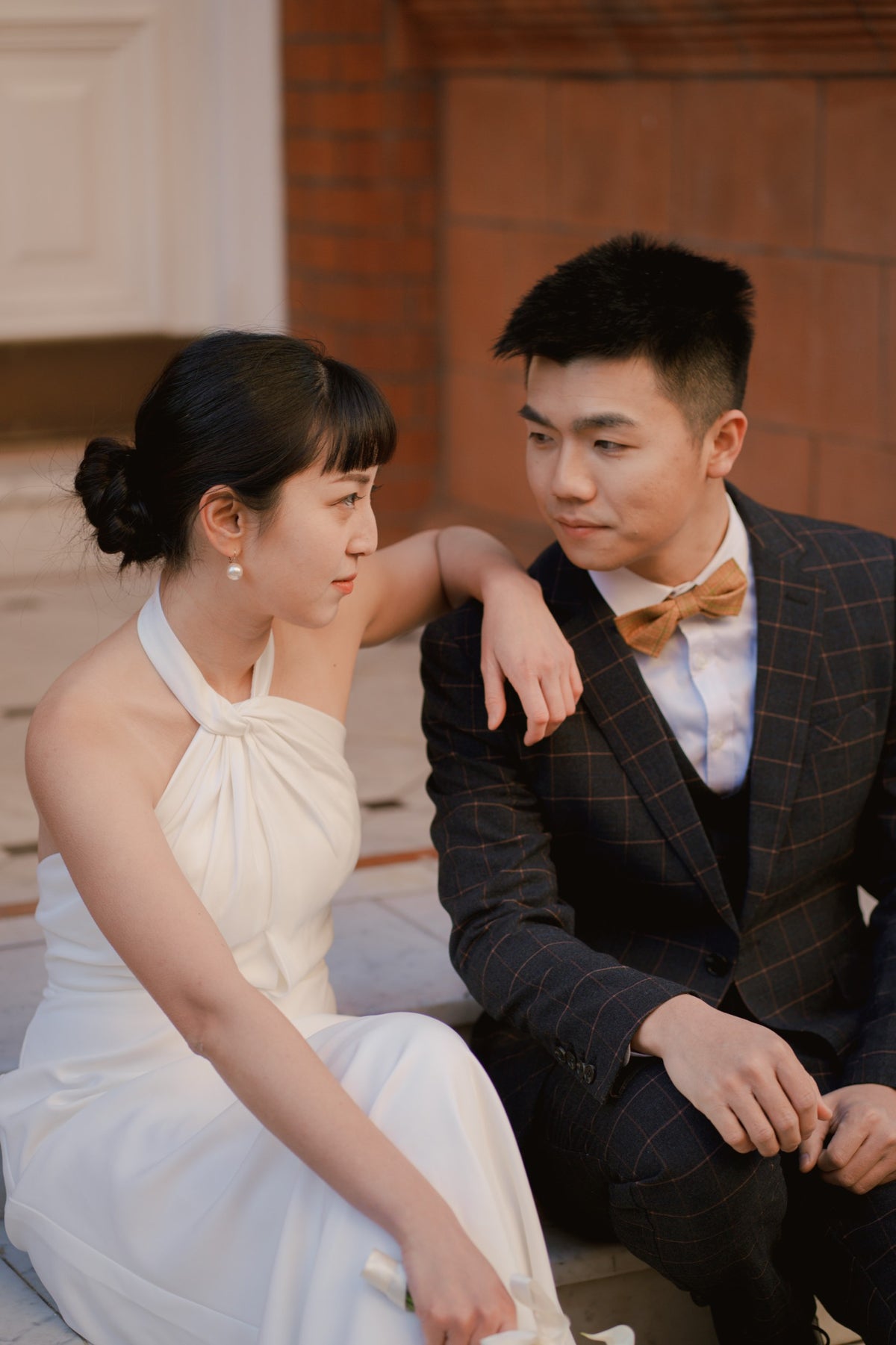 A couple in formal wedding attire sitting together outdoors. The woman wears a sleeveless white halter gown, and the man wears a dark plaid suit jacket with a brown bow tie over a white shirt.