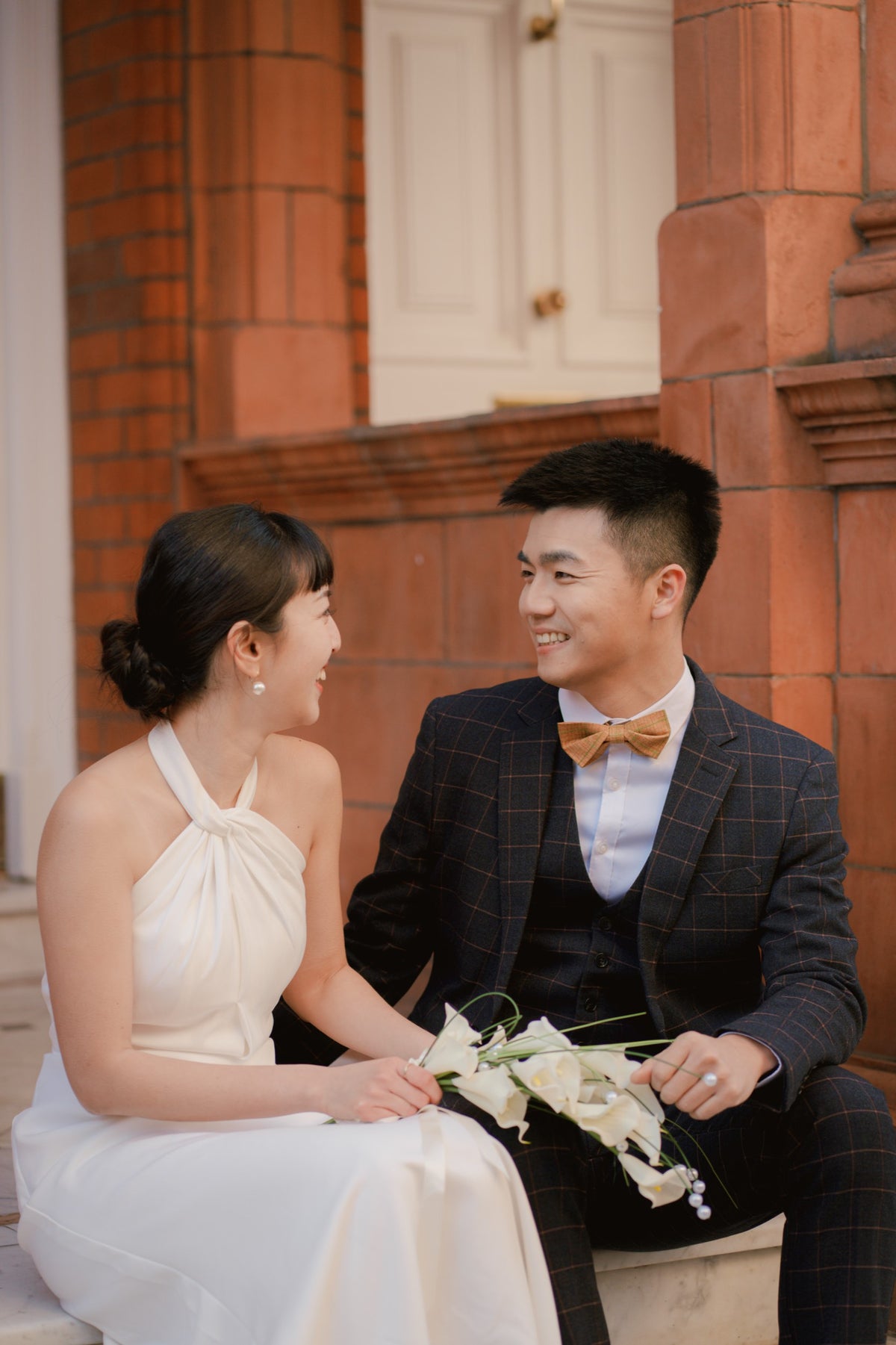 A bride in a white halter wedding dress and a groom in a dark plaid suit with a tan bow tie sit together on stone steps in front of a red brick building, holding a bouquet of white flowers.