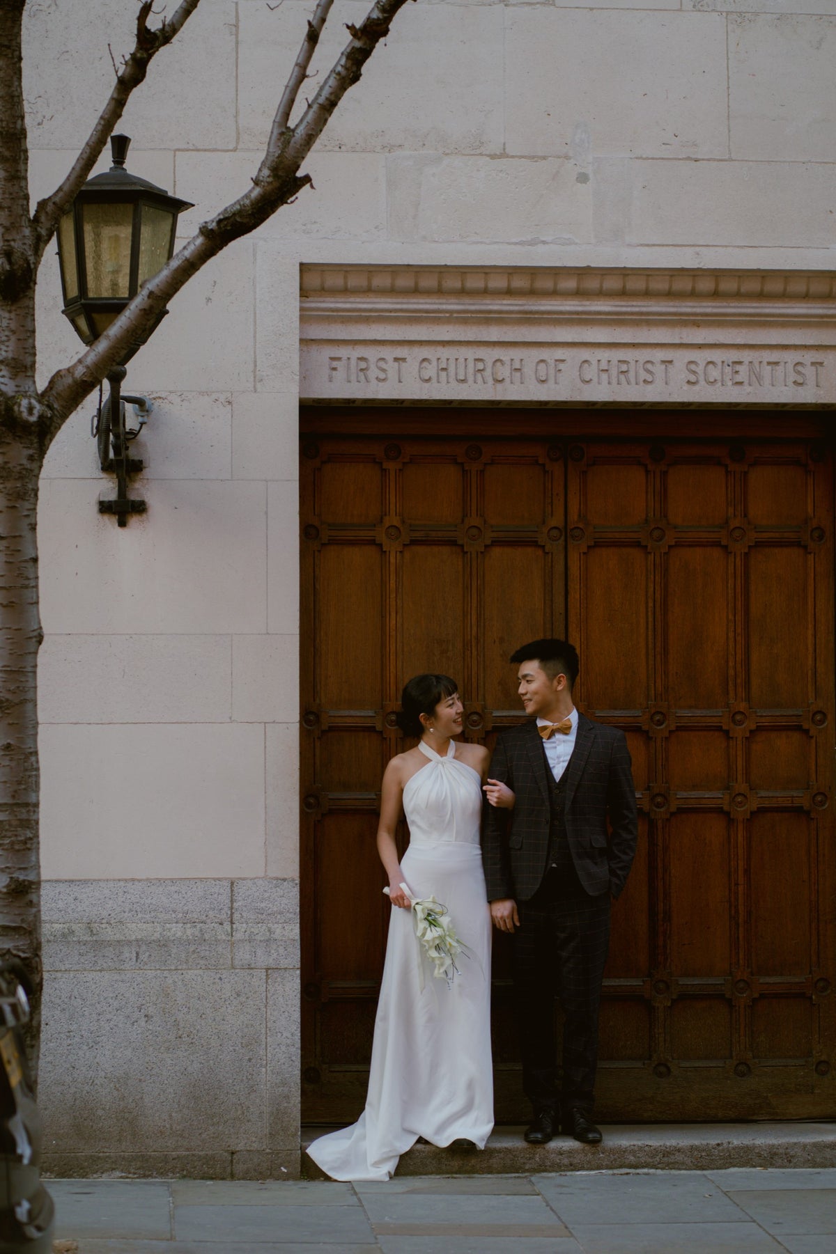 A bride in a white sleeveless wedding gown and a groom in a dark suit stand in front of a large wooden door labeled 'FIRST CHURCH OF CHRIST SCIENTIST' on a stone building facade.