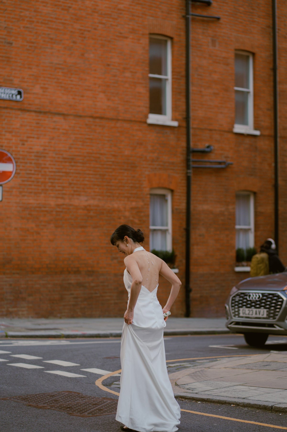 A woman in a sleeveless white wedding gown stands on a city street in front of a red brick building, holding the skirt of the dress out to show its drape and length.