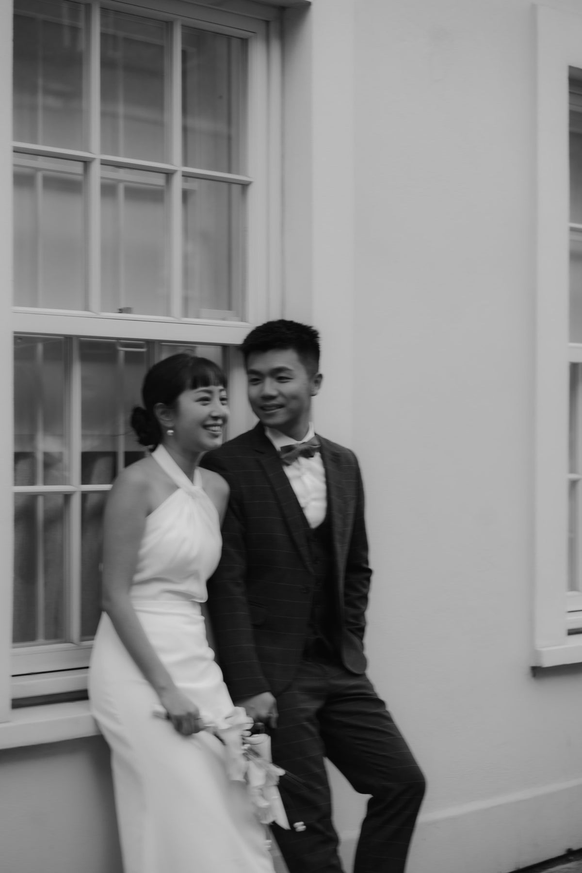 A black and white photograph of a bride and groom posing together against a white stucco wall with large windows.
