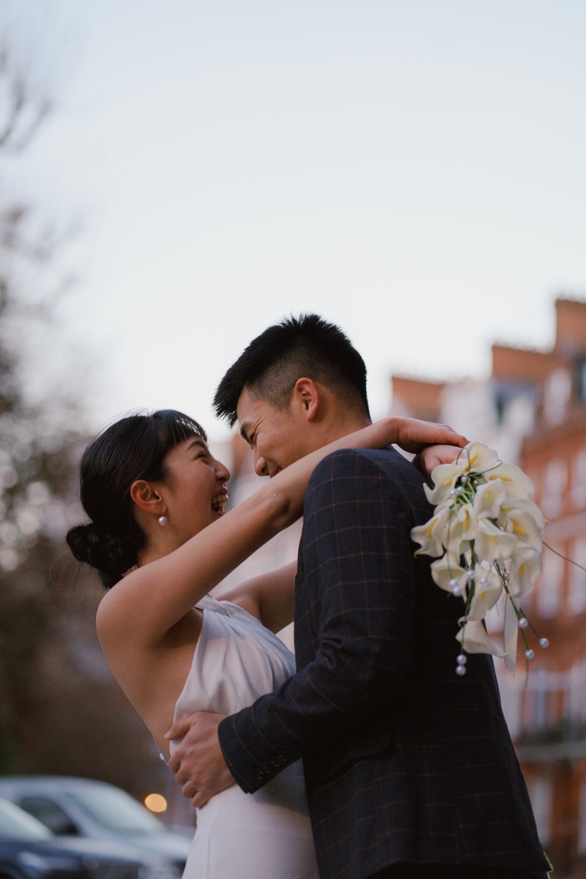 A bride in a white wedding dress and a groom in a dark plaid suit embrace and kiss outdoors, with a bouquet of white flowers and a softly blurred city background.