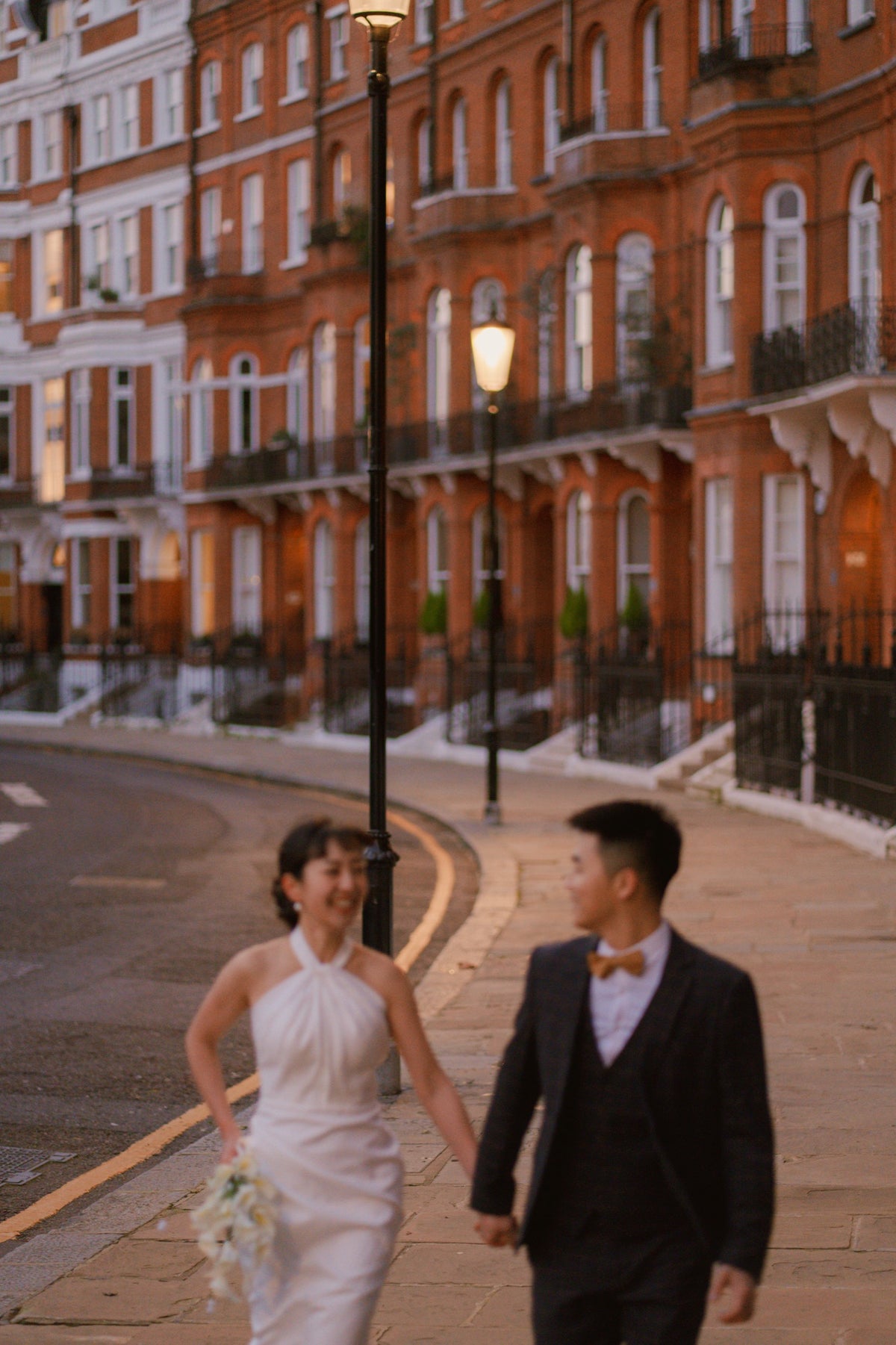 A bride in a white sleeveless wedding dress and a groom in a dark suit walking hand in hand along a city street lined with warm orange brick townhouses at dusk.