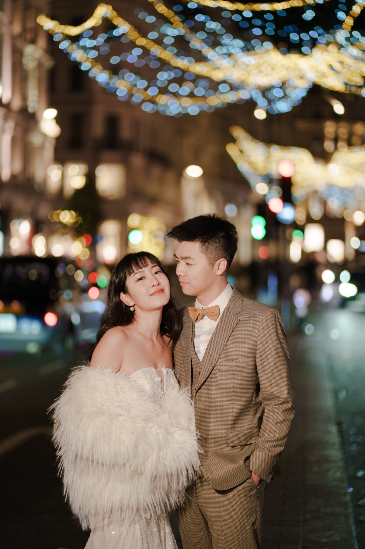 A couple posing on a city street at night decorated with festive bokeh lights. The woman is wearing a strapless white gown with a fluffy feathered shawl, and the man is wearing a brown plaid suit with a bow tie.