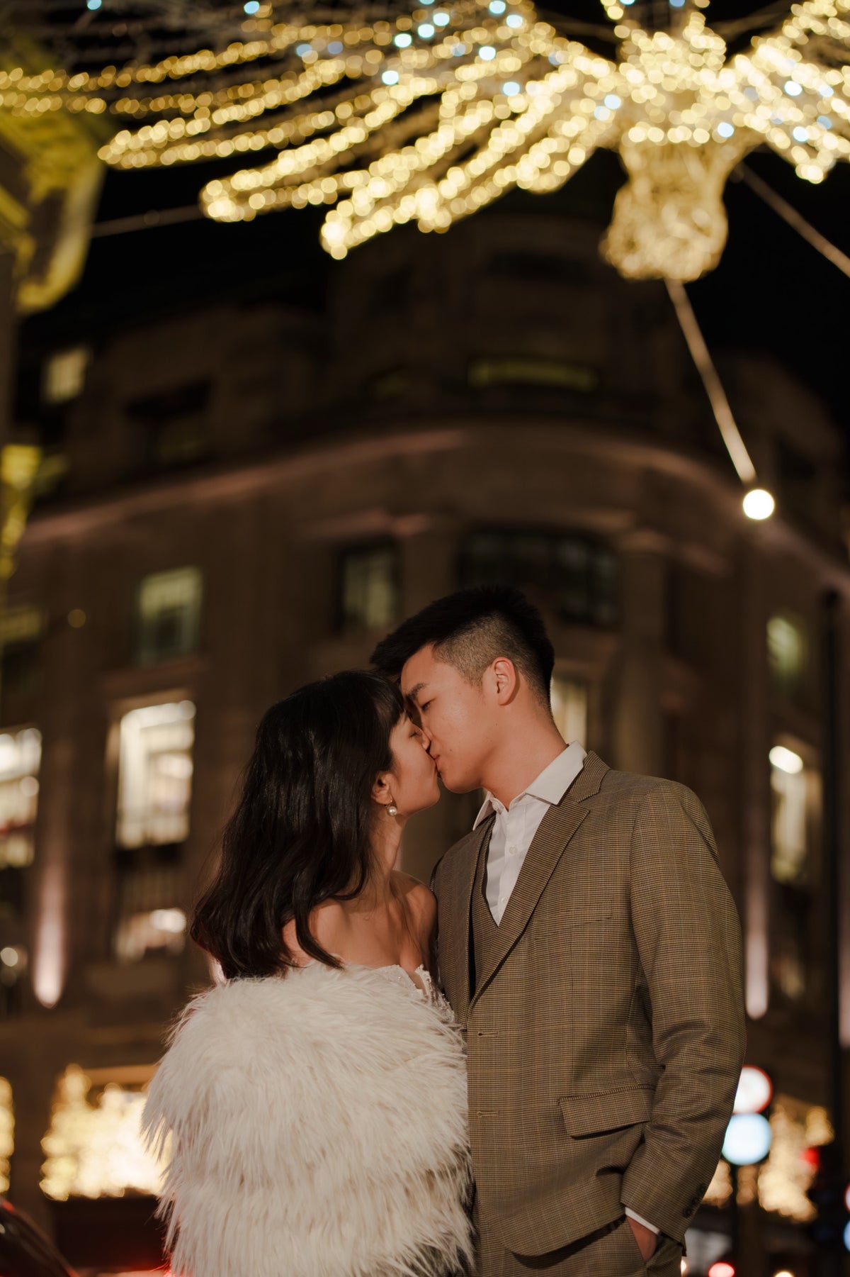 A couple embracing and kissing in an outdoor city setting at night, decorated with warm golden fairy lights. The woman wears a fluffy white faux-fur jacket, and the man wears a brown plaid suit jacket over a white shirt.
