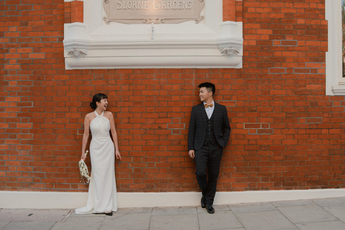 A bride in a white halter-neck wedding gown and a groom in a dark suit with a bow tie stand on a sidewalk in front of a red brick wall with a white decorative plaque.