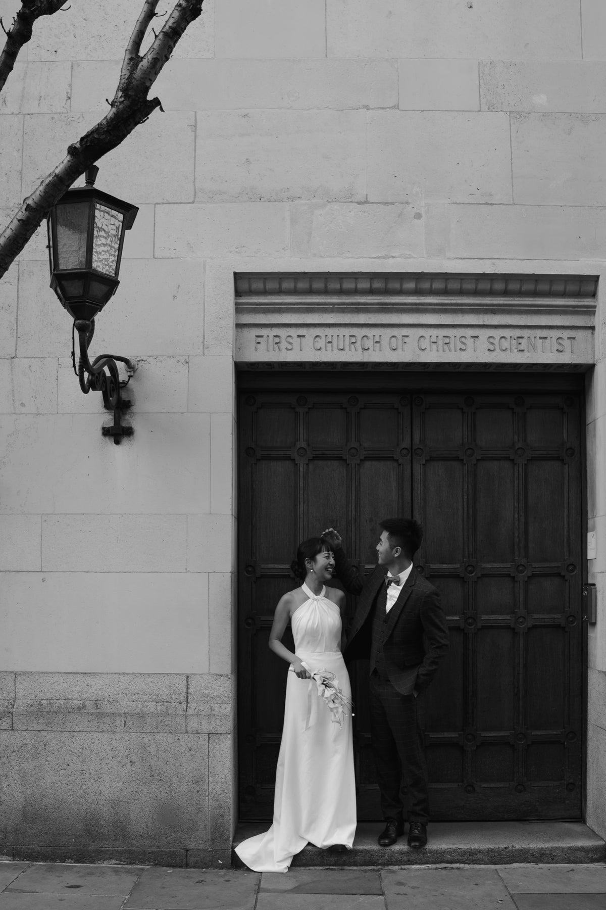 Black and white wedding photograph of a bride in a sleeveless white gown and a groom in a dark suit standing in front of a large dark wooden door on a stone building, with the inscription 'FIRST CHURCH OF CHRIST SCIENTIST' above the door.