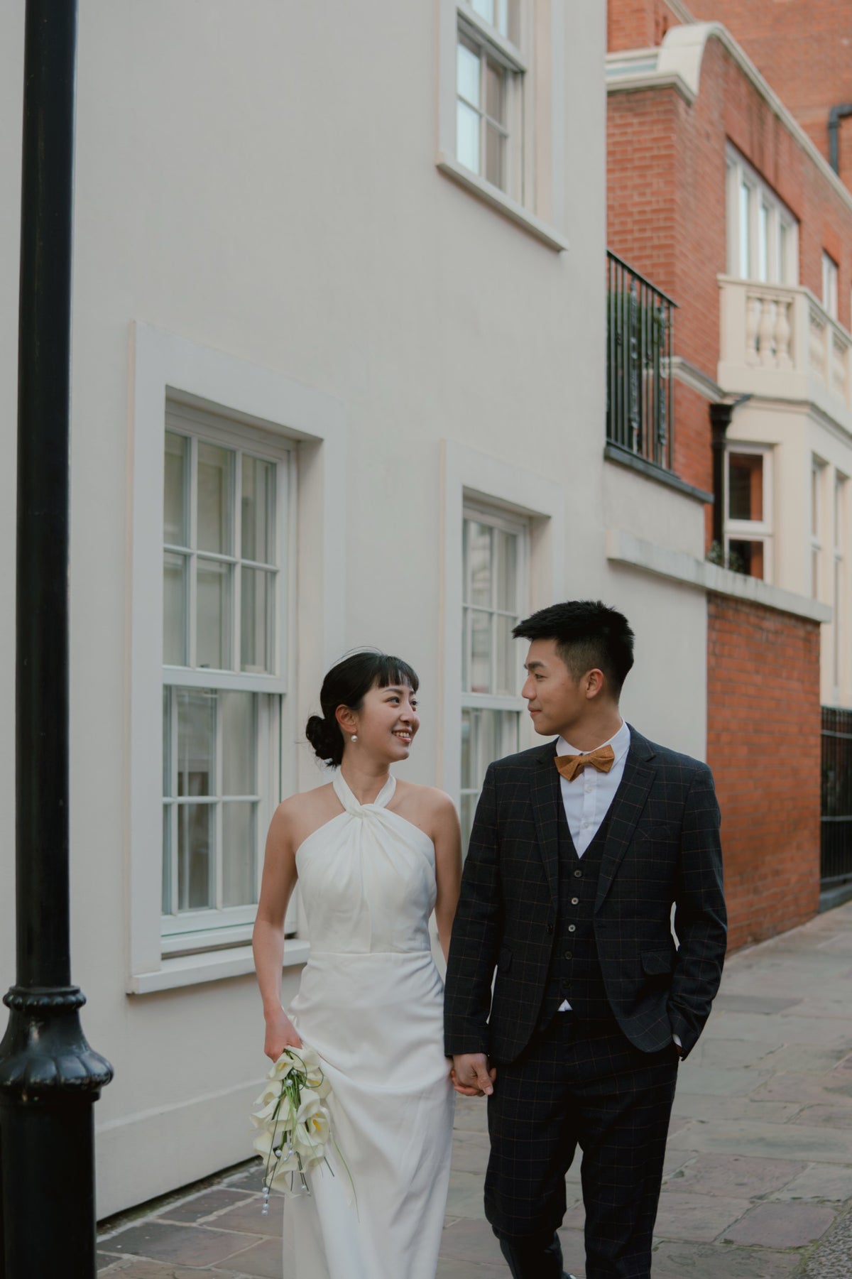 A bride in a white halter-neck wedding gown holding a bouquet of white flowers walks hand-in-hand with a groom in a dark plaid suit and a yellow bow tie down a cobblestone street lined with white and red brick buildings.