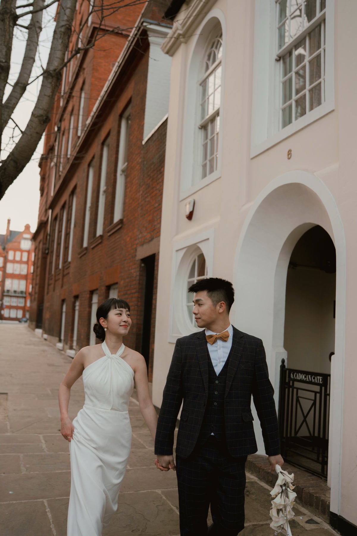 A bride in a white halter-neck wedding gown and a groom in a dark gray plaid suit with a yellow bow tie walk hand-in-hand down a cobblestone street lined with historic brick and stucco buildings.